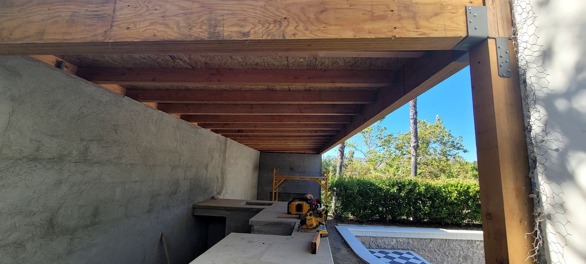 Construction site showing a wooden roof frame with exposed beams over a grey concrete wall and outdoor work area.