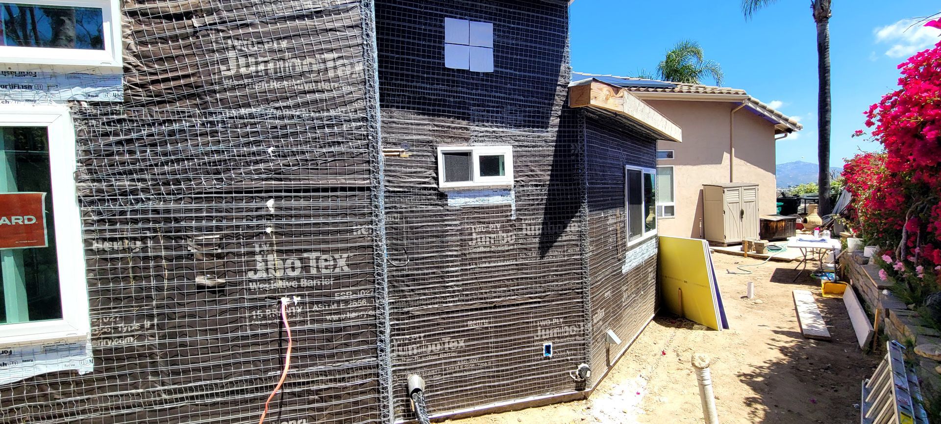 An exterior view of a house under construction, covered in black weather-resistant wrap with metal lath for stucco.
