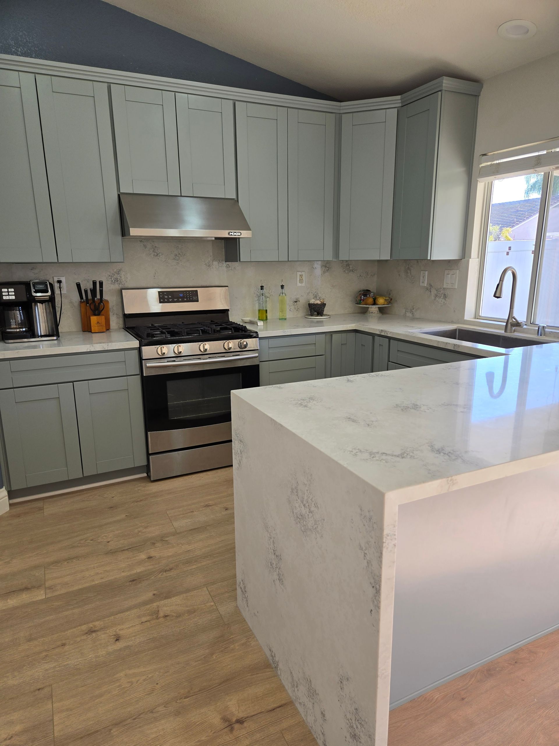 A kitchen with light gray cabinets, white marble-patterned countertops, a stainless steel stove, and light wood flooring.
