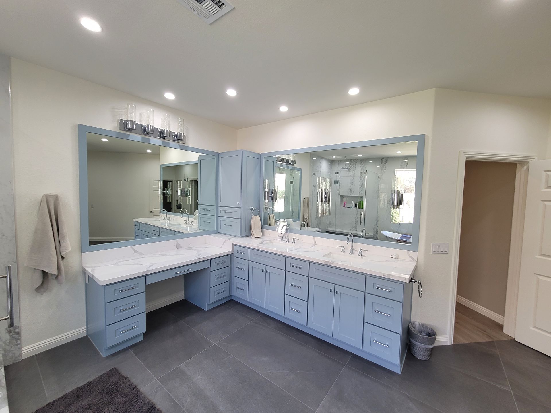 A bright bathroom with light blue L-shaped cabinets, white countertops, dual mirrors, and grey tile flooring.