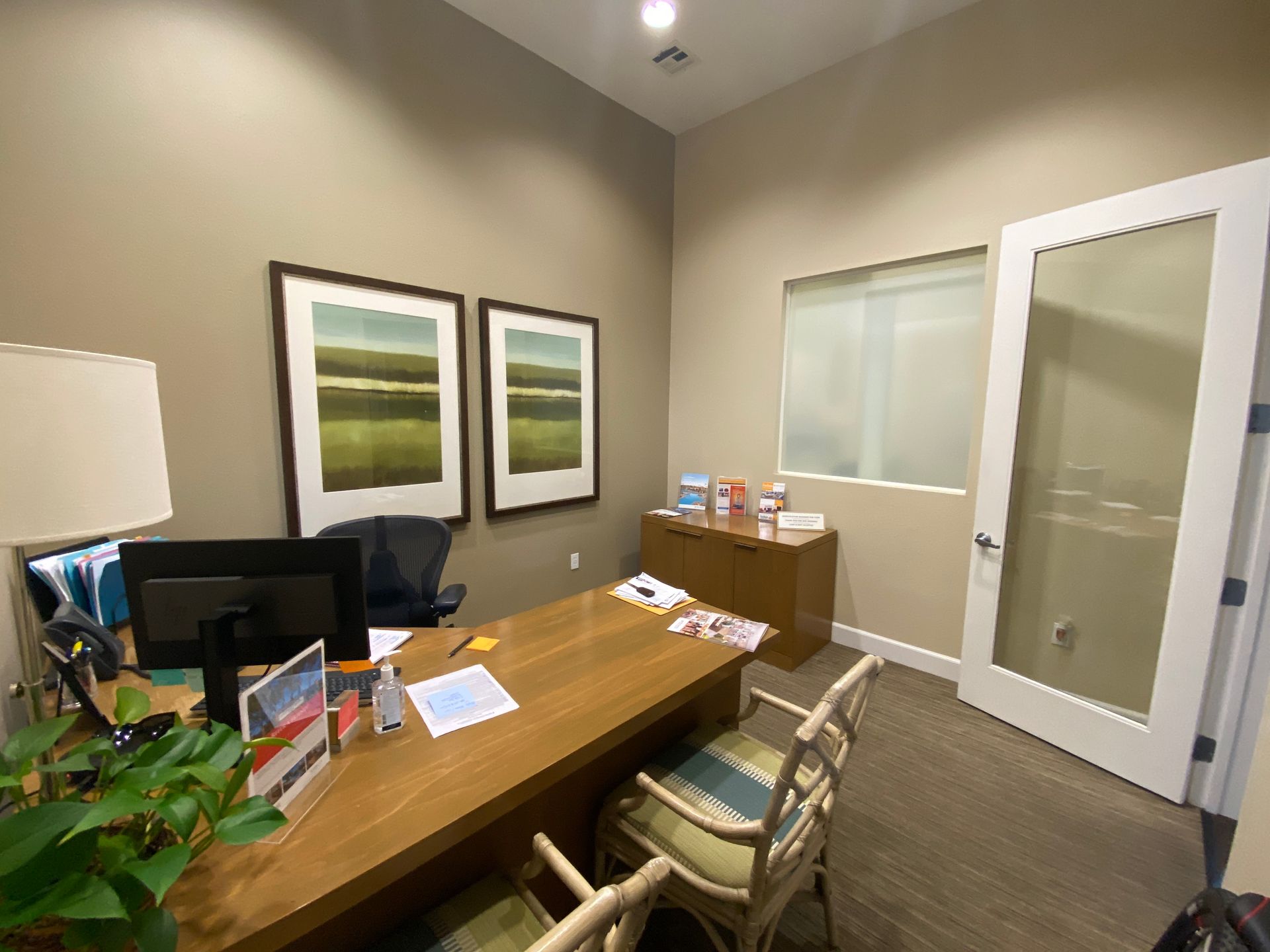 A small office with a wooden desk, computer, two framed landscape prints on a tan wall, a chair, and a glass-paneled door.