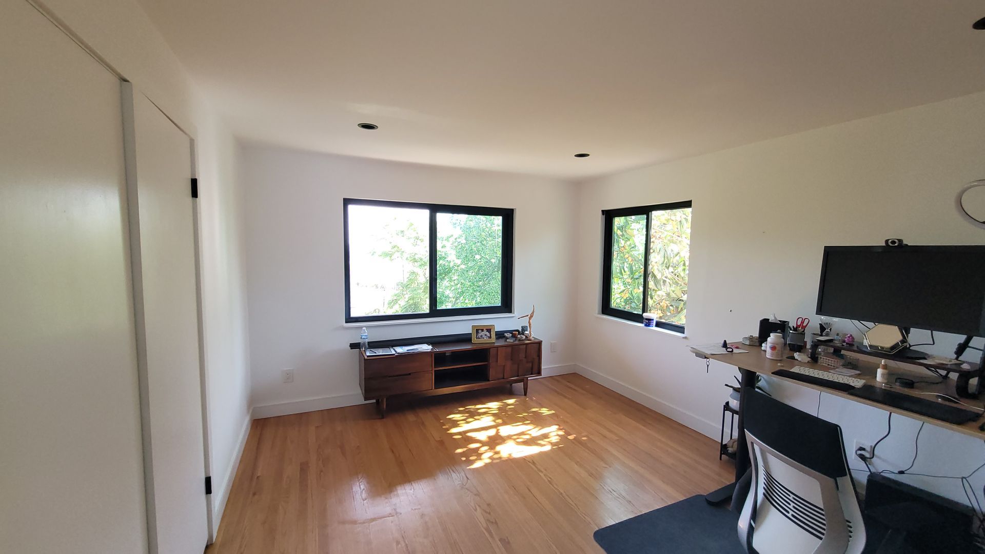 A home office with light hardwood floors, white walls, two windows with black frames, a wood media console, and a desk.