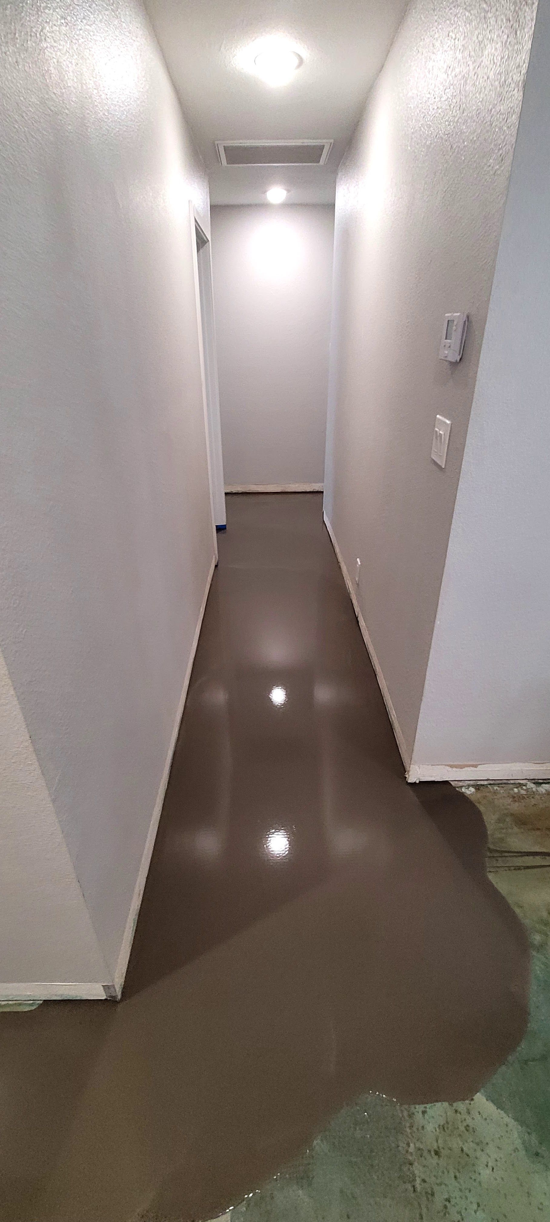 A hallway with freshly poured, wet self-leveling concrete covering the floor, transitioning to bare subflooring.