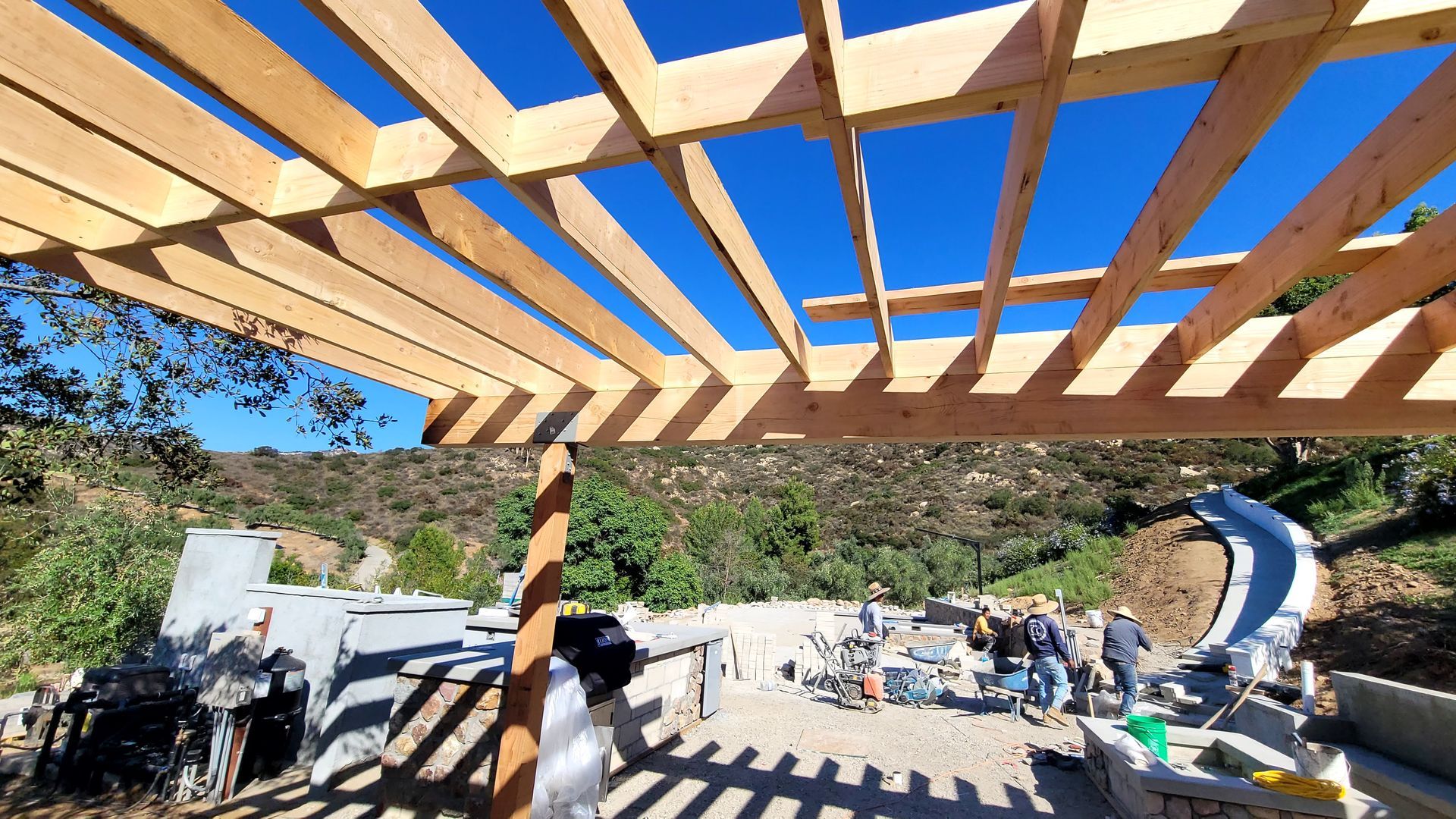 A wooden pergola frame stands over a construction site with workers and concrete structures on a sunny hillside.