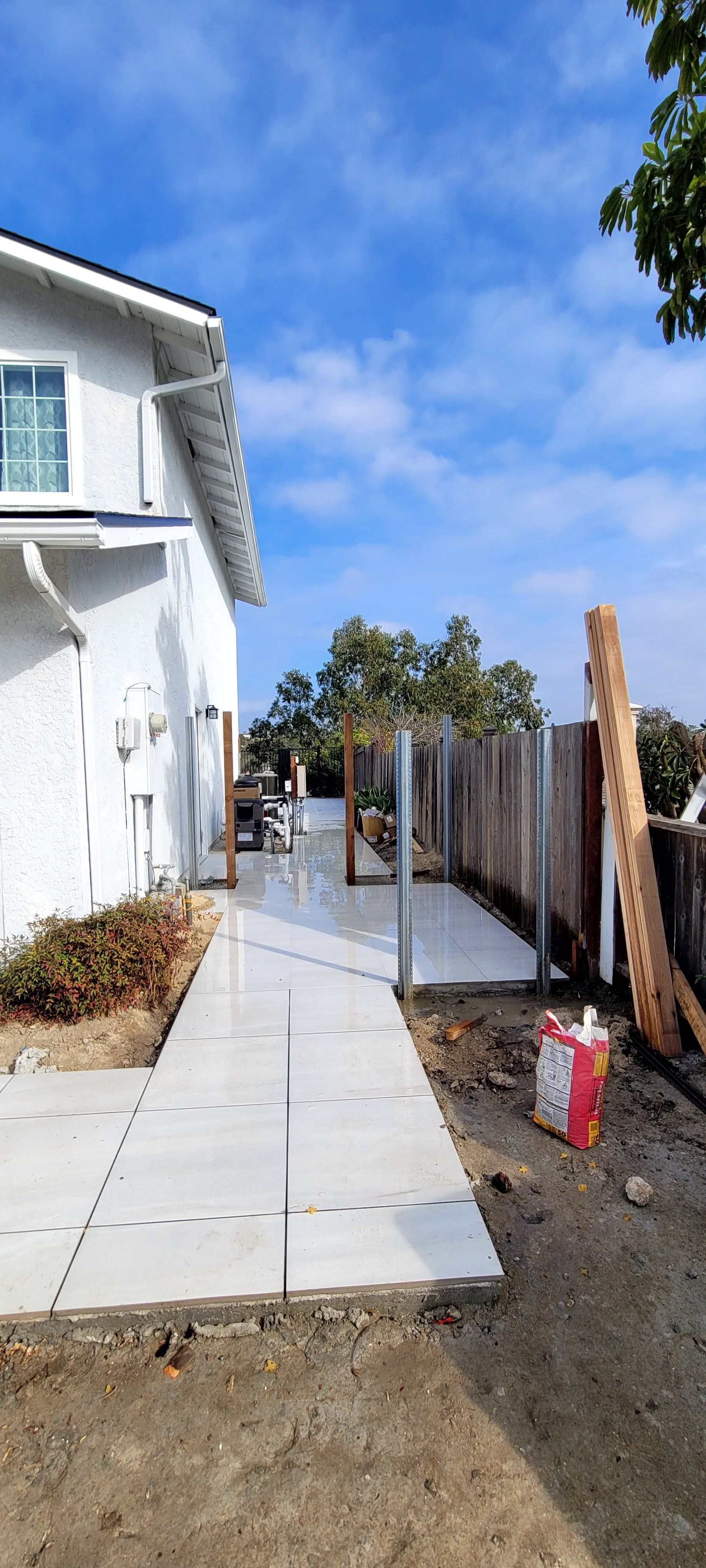 White square tiles are being laid along a walkway next to a white house and a wooden fence under a sunny blue sky.