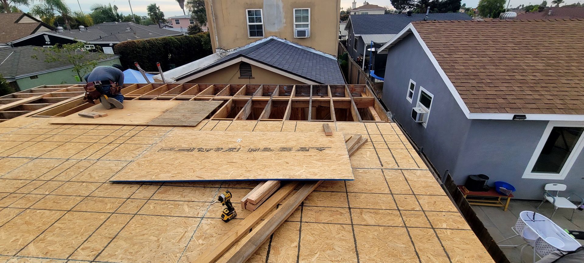 A worker installs plywood sheeting on a roof under construction, adjacent to a gray house and a blue-roofed building.