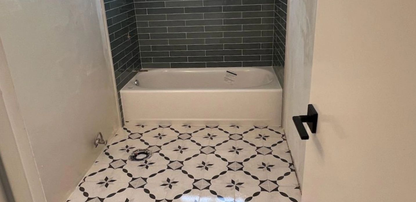 A bathroom featuring a white bathtub, dark gray subway tile shower surround, and patterned black-and-white floor tiles.