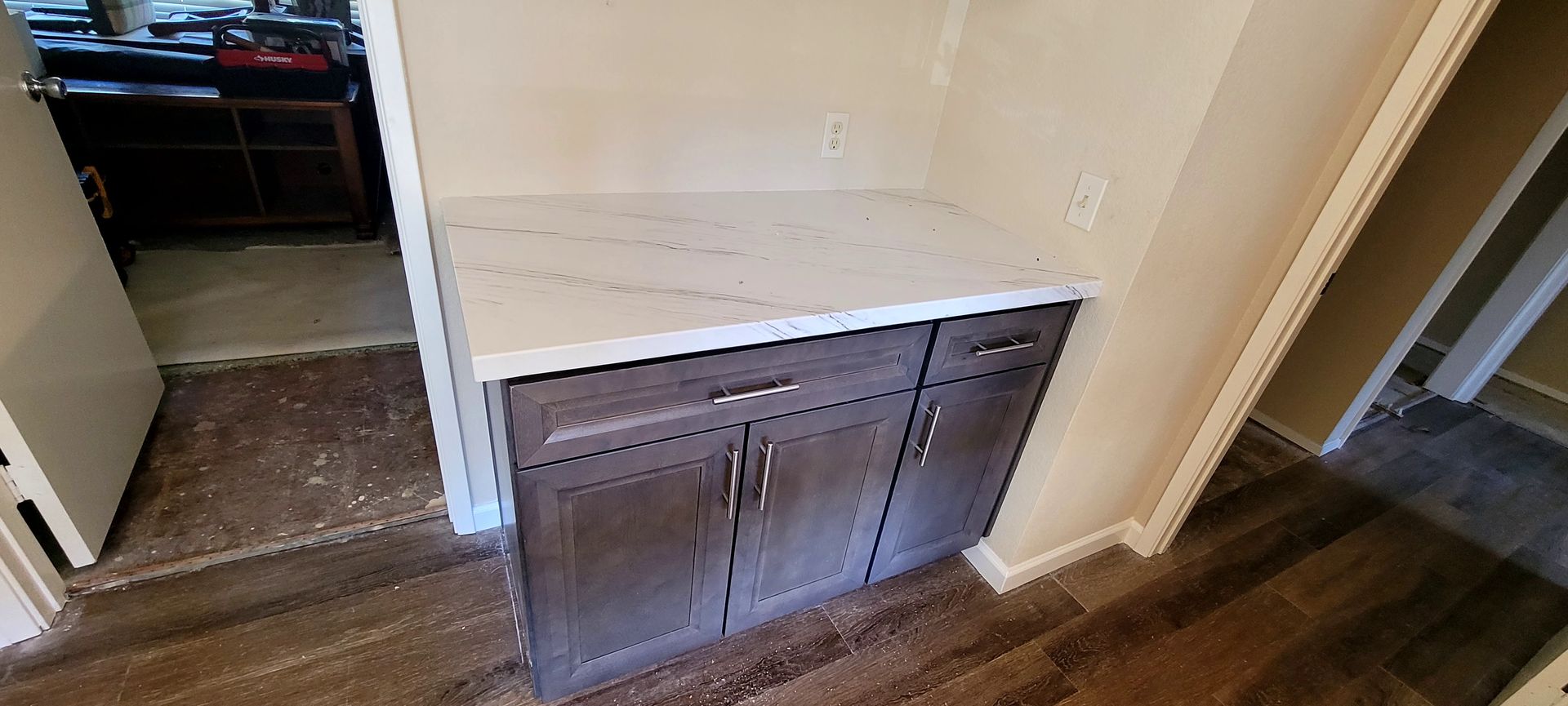 A white marble countertop sits on dark gray kitchen cabinets in a room with wood-look flooring and an adjacent doorway.