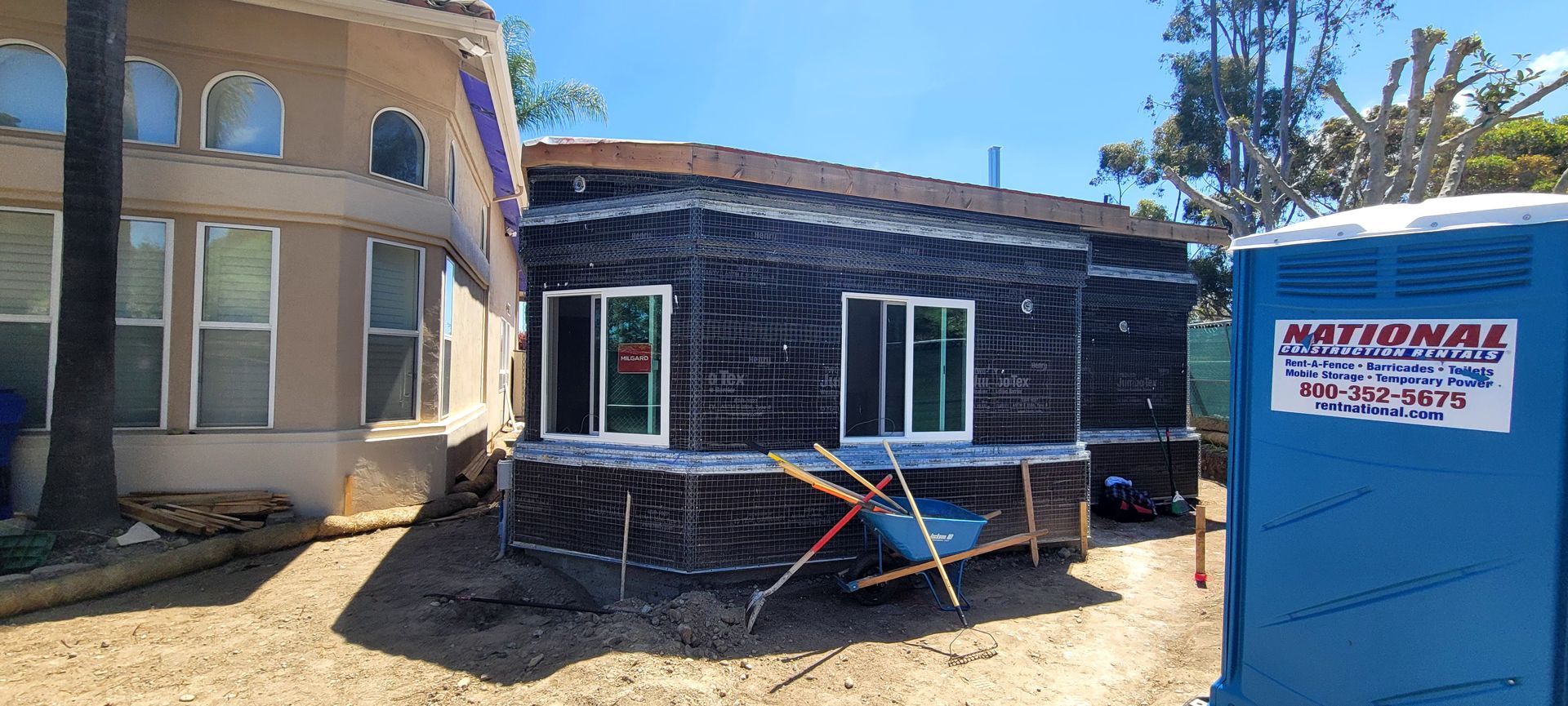 A house under construction with black building paper siding next to a tan house and a blue portable toilet on a dirt lot.