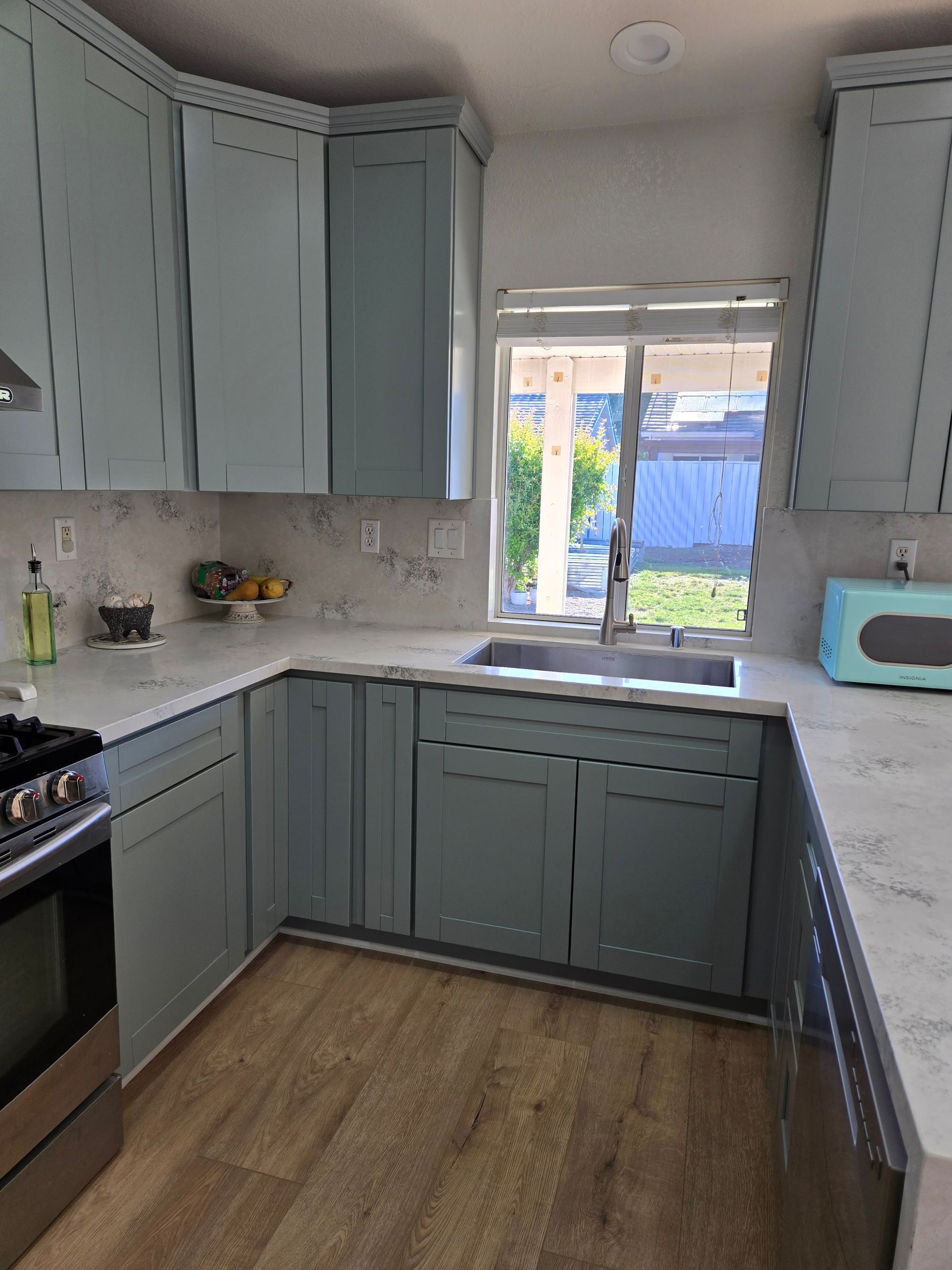 A kitchen with light blue cabinets, white marble-patterned countertops, a stainless steel sink, and wood-look flooring.