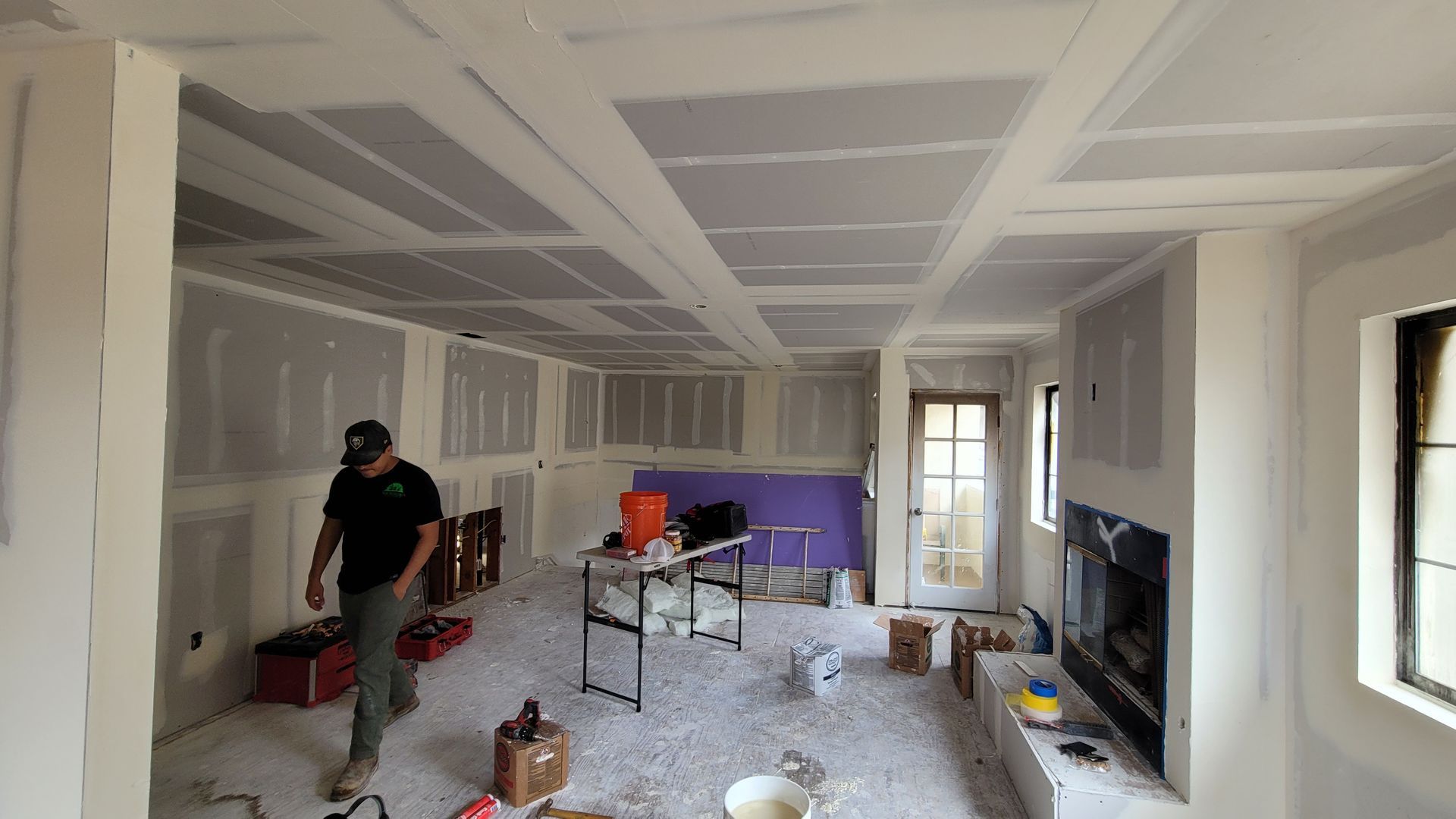 A worker walks through a room under renovation with drywall installed on walls and ceiling, a fireplace, and supplies.