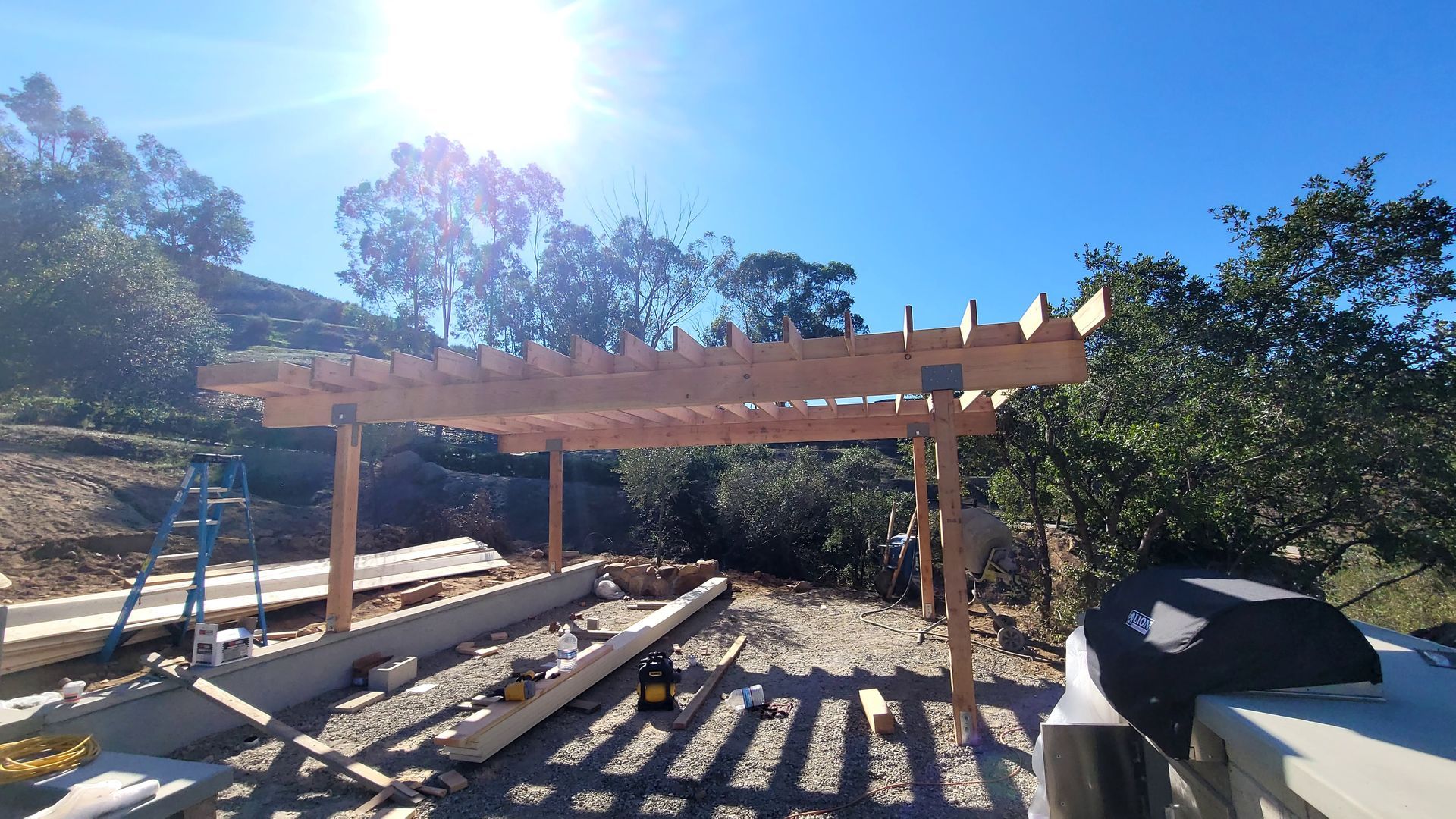 A wooden pergola frame under construction in a sunny, outdoor backyard setting.
