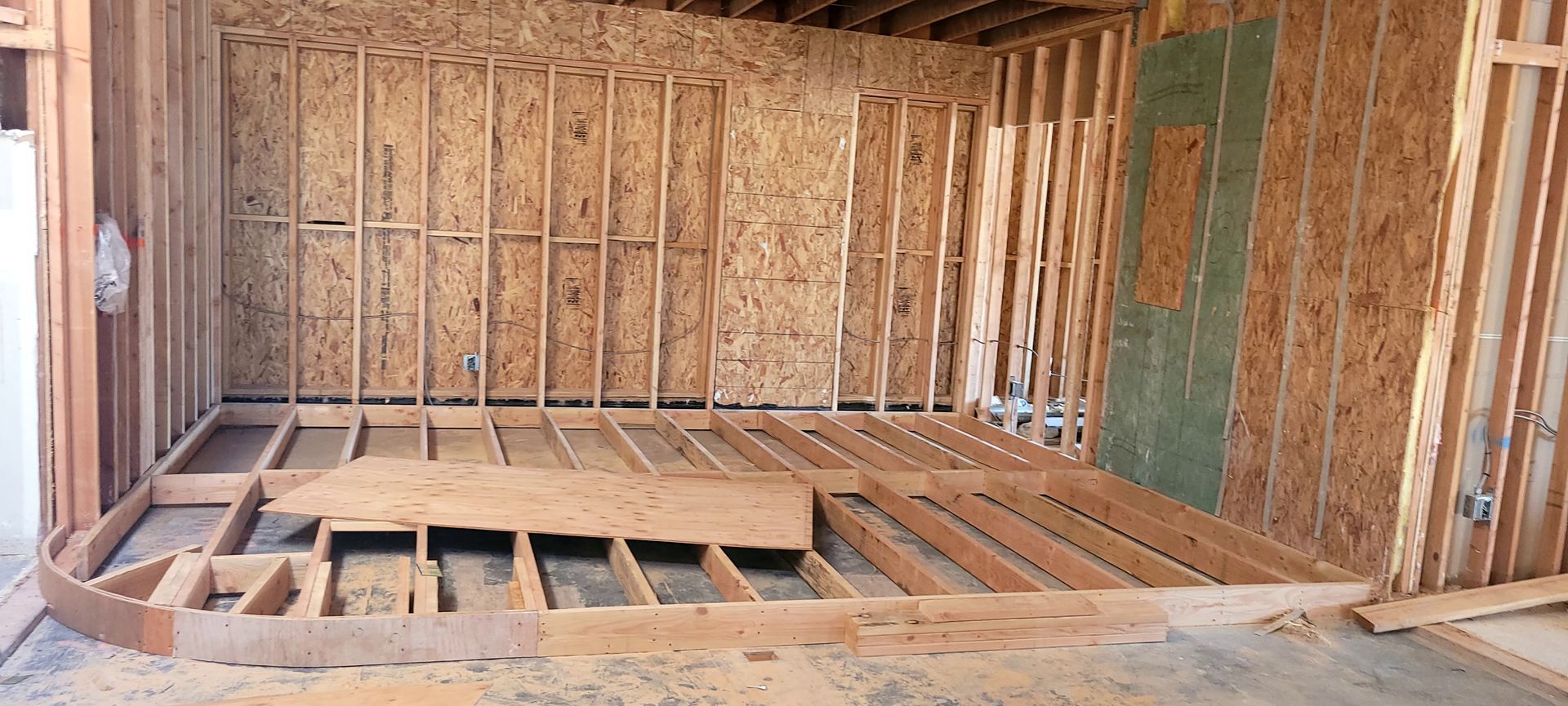 Framed interior of a room under construction, showing wooden floor joists, wall studs, and a section of green wallboard.