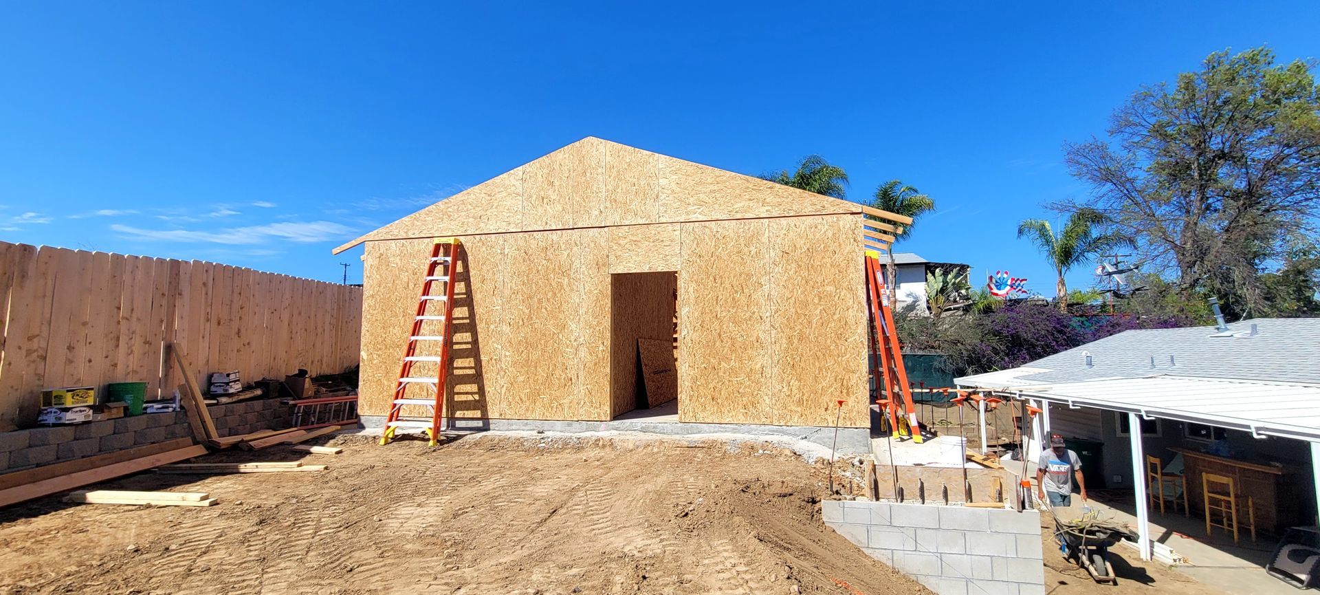 Construction of a small, wood-sheathed building under a clear blue sky, featuring ladders leaned against the exterior.
