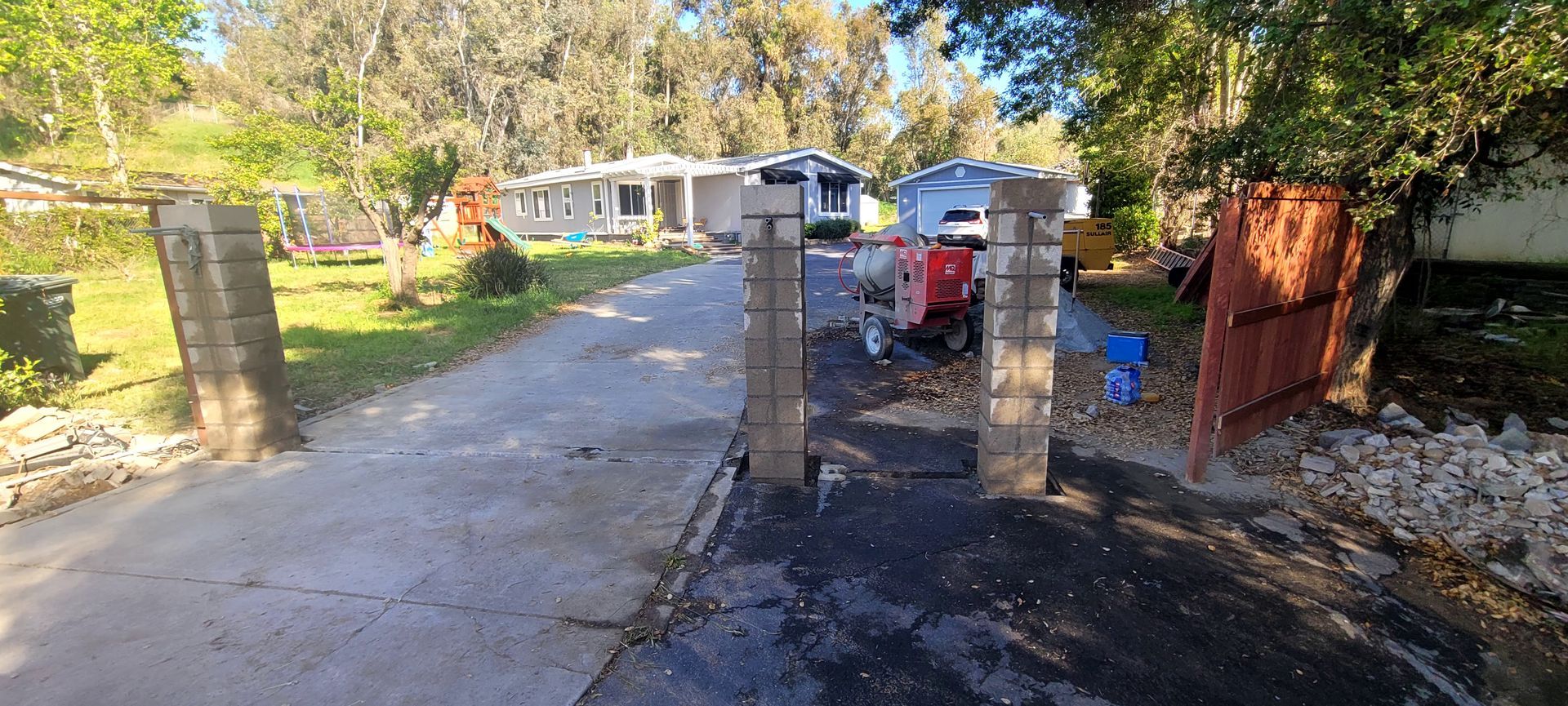 Three masonry gate pillars stand on a paved driveway in front of a residential house surrounded by trees.