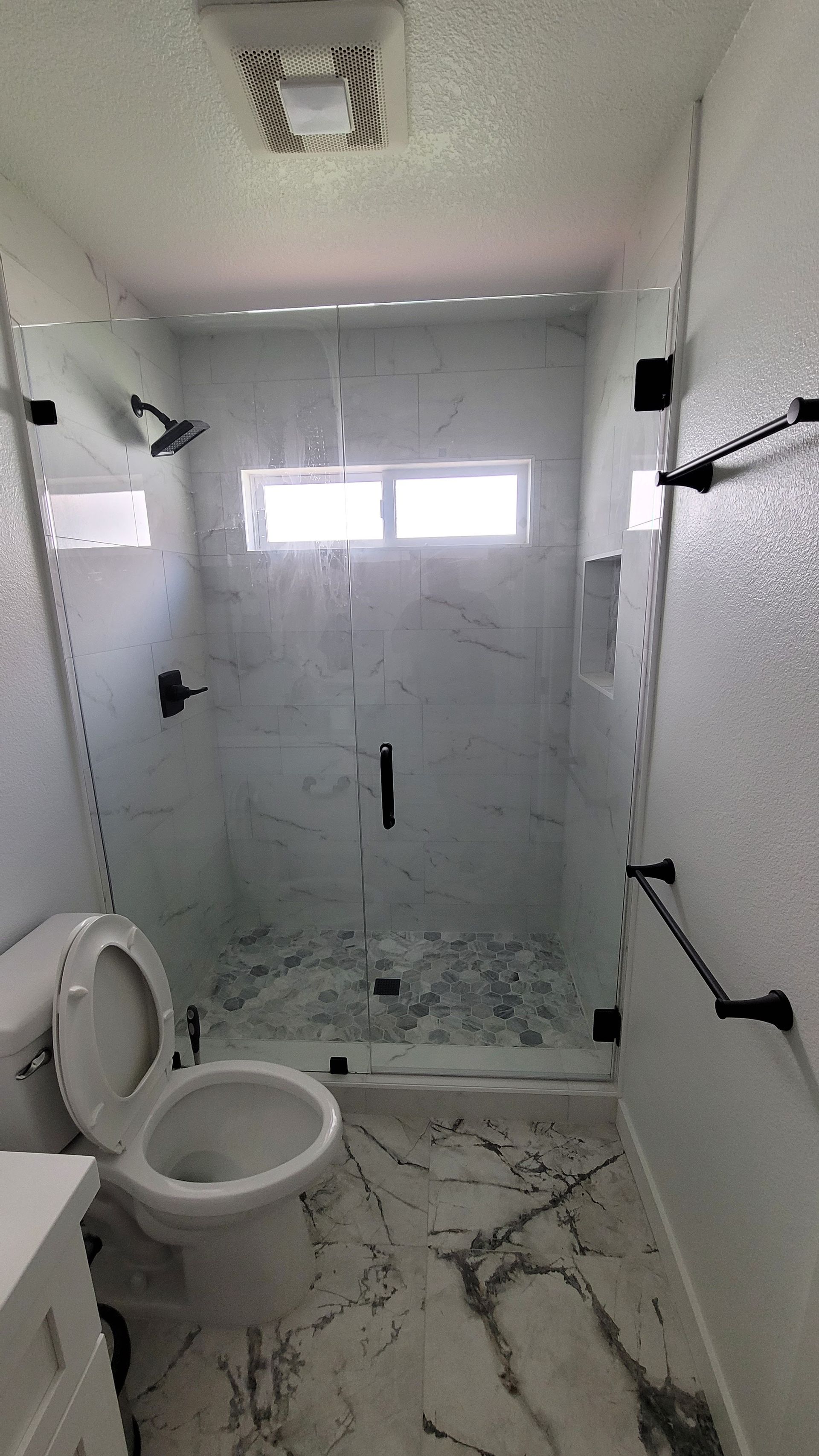 A modern bathroom with white marble-patterned flooring, a toilet, and a glass-enclosed shower with black hardware.