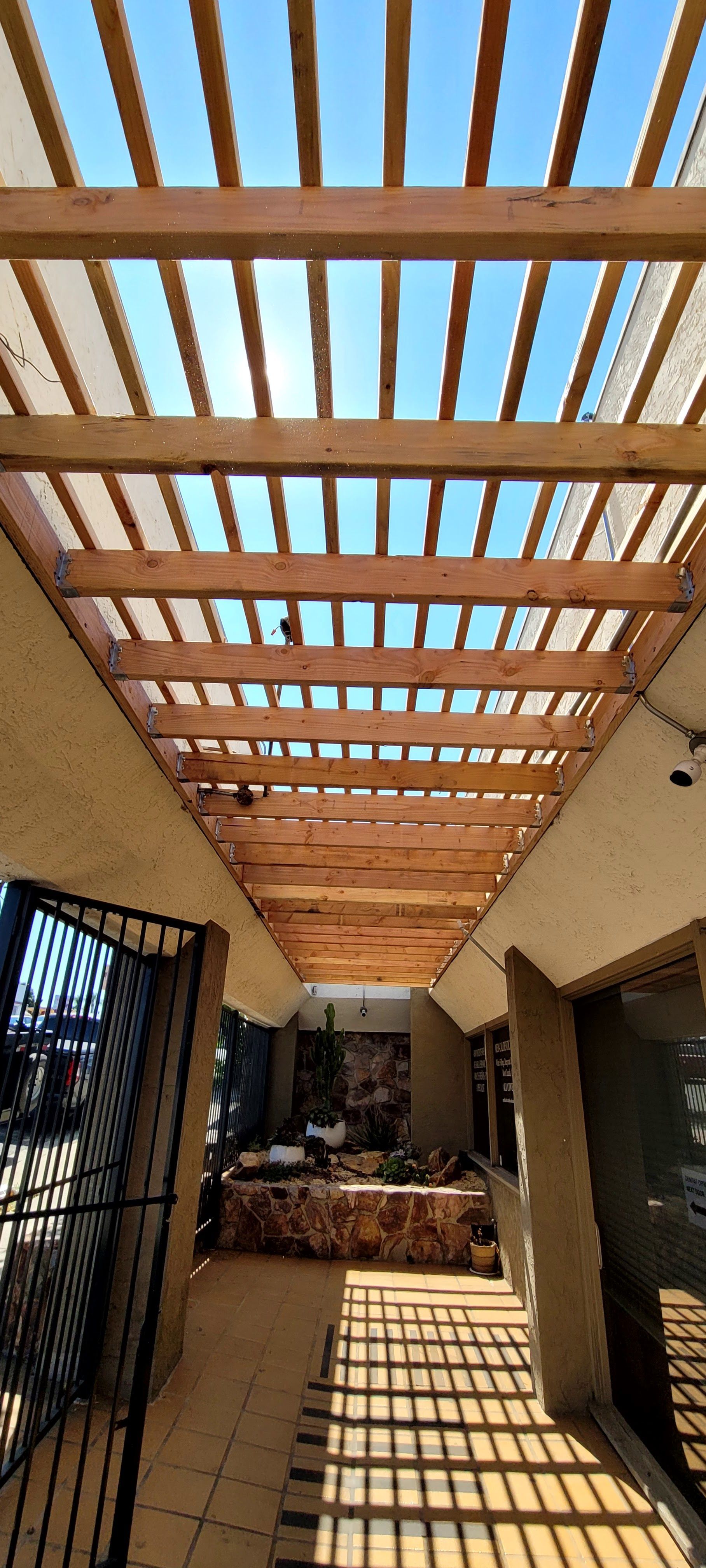 A view through a wooden pergola roof, casting linear shadows across a paved outdoor walkway leading to a storefront area.