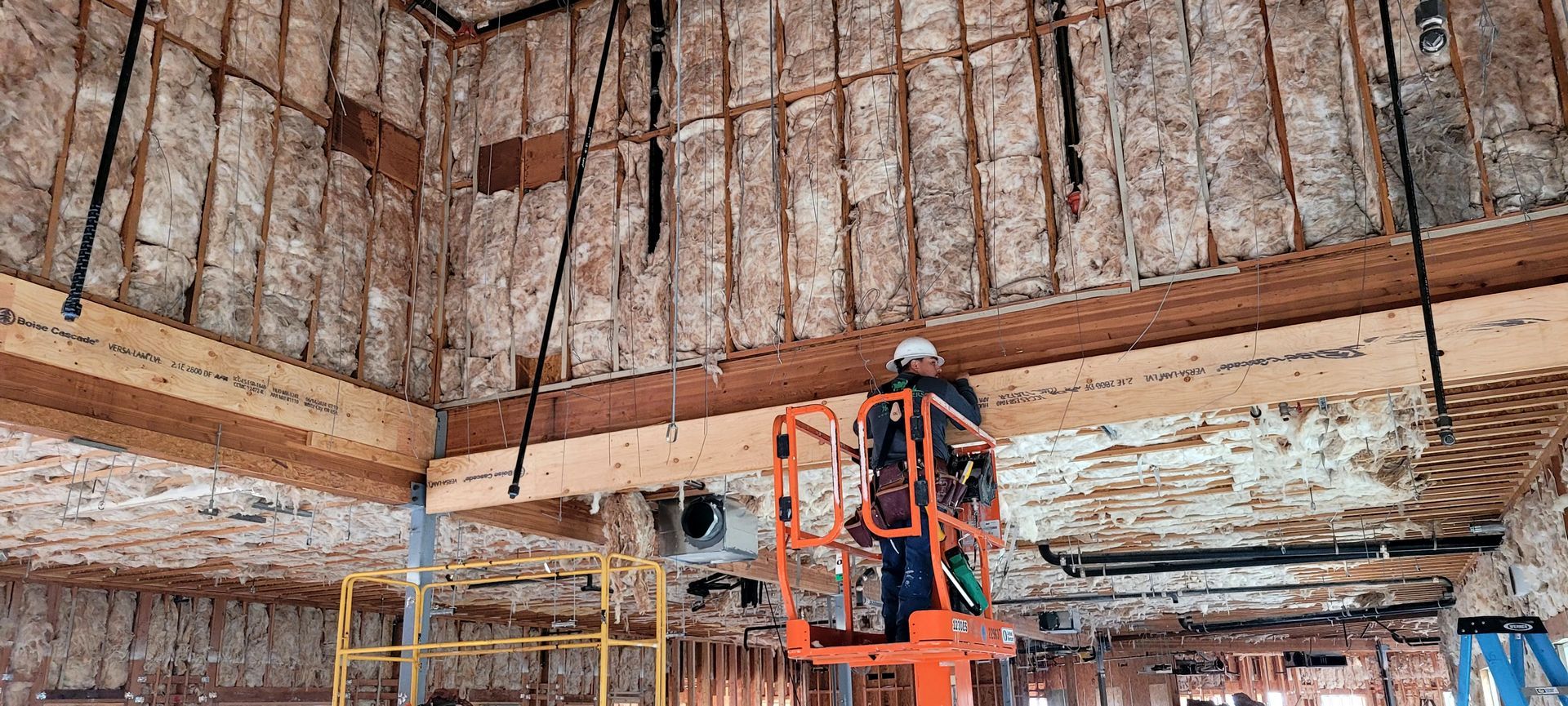A worker in a safety helmet stands on an orange scissor lift installing insulation in the rafters of a building frame.