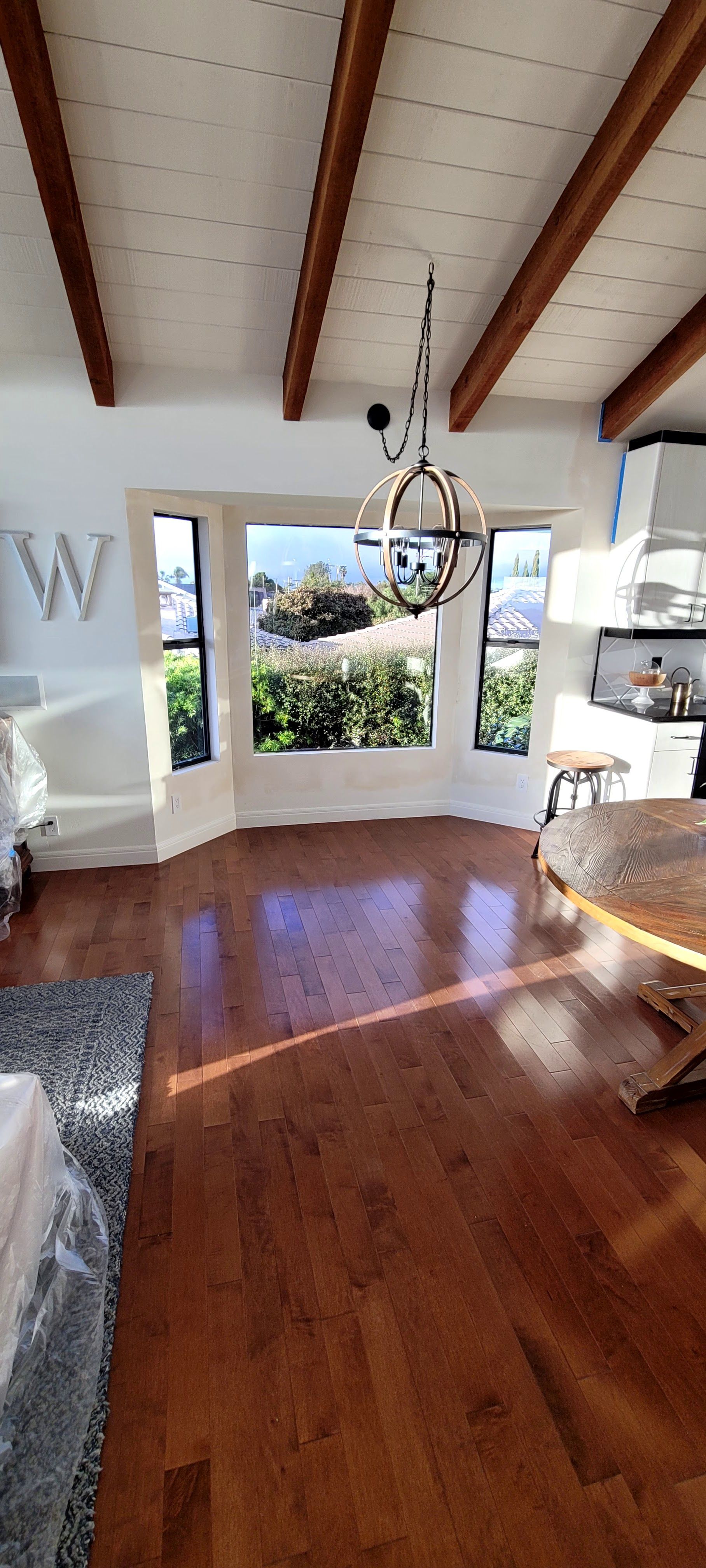 A sunny dining area with hardwood floors, a large bay window, wooden ceiling beams, and a round chandelier.