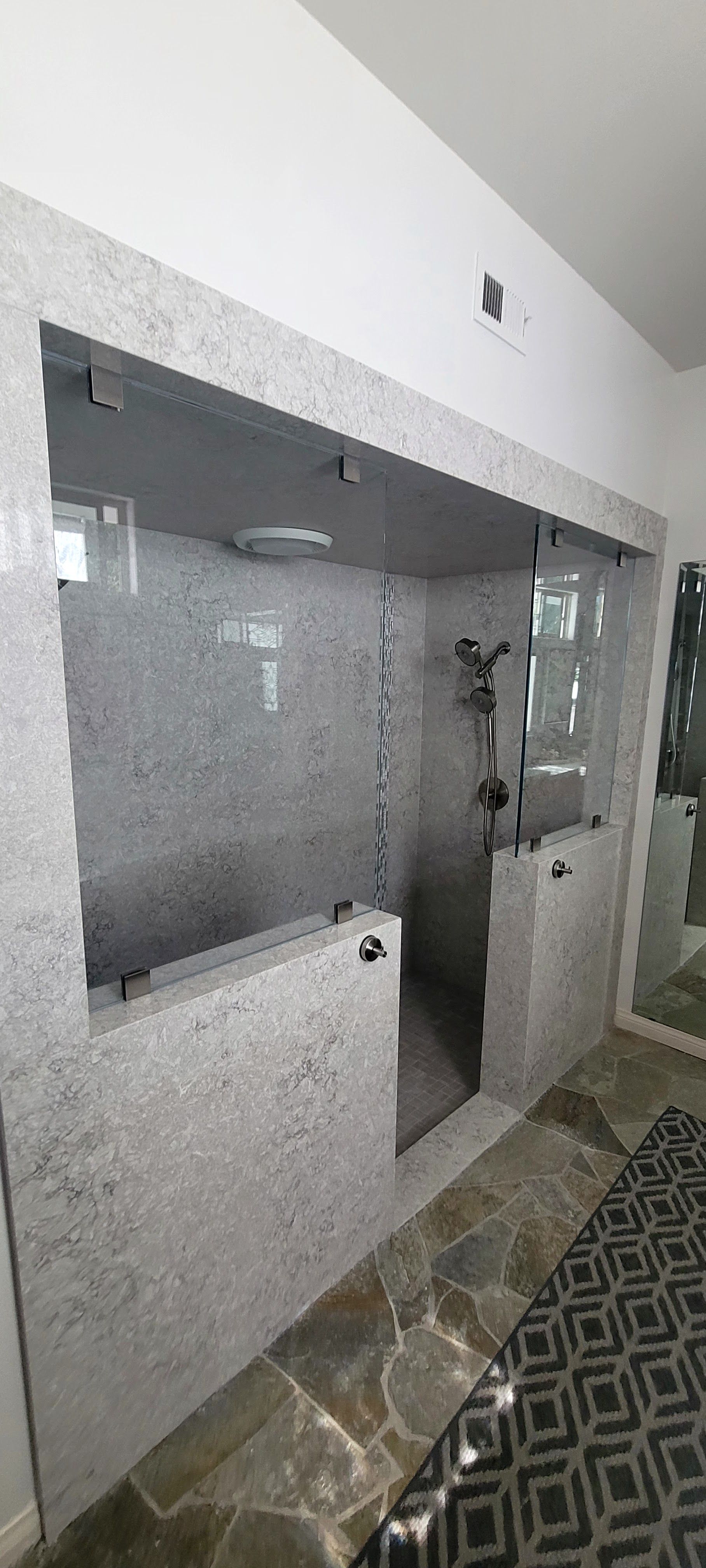 A modern walk-in shower with light grey stone walls, glass panels, and a patterned rug on the floor.