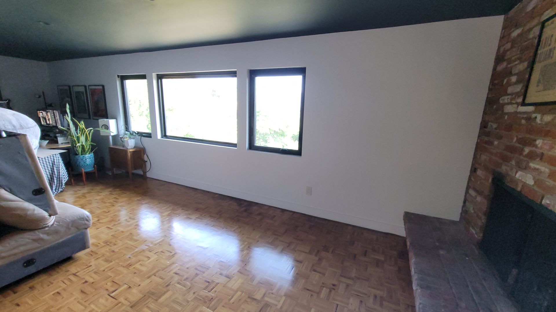 A room with parquet wood floors, white walls, a dark ceiling, three windows, and a brick fireplace.