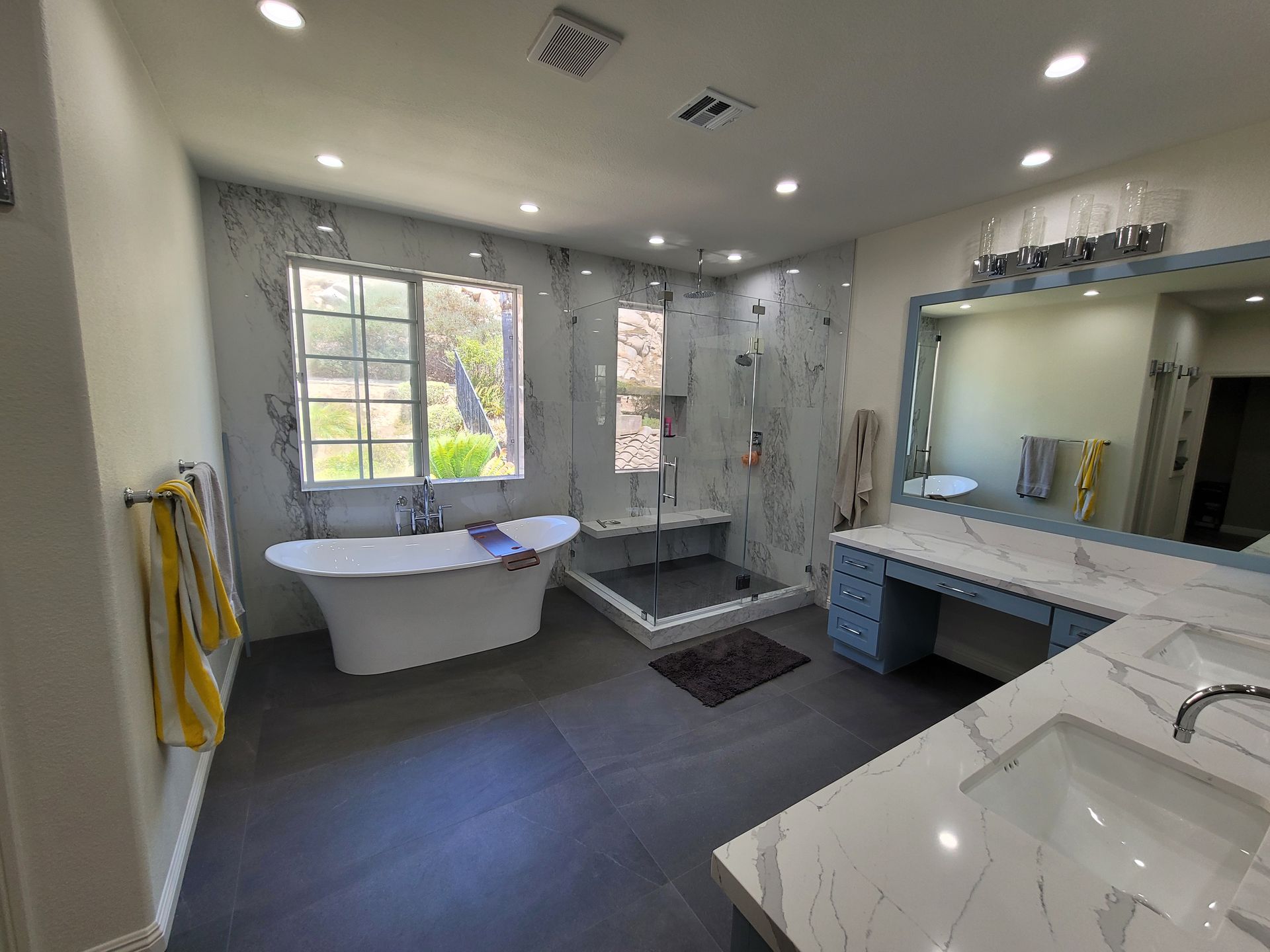 A bright bathroom with a freestanding tub, glass walk-in shower, dual marble-topped vanities, and gray tiled floors.