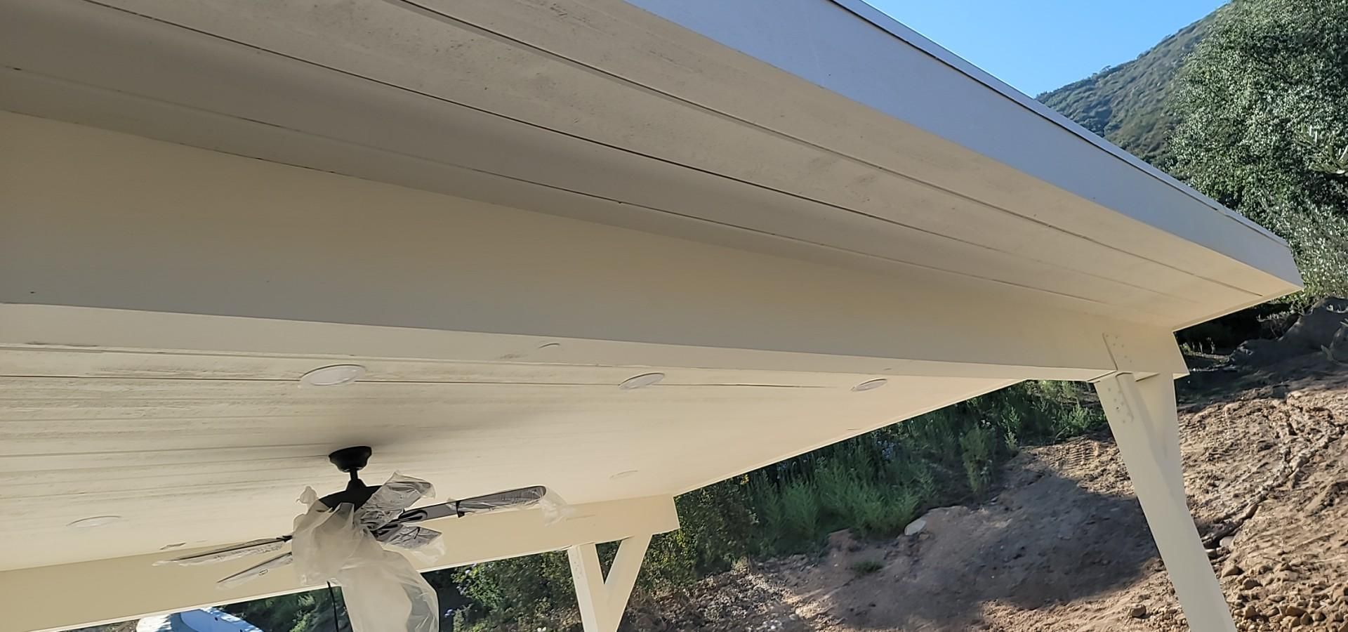 A low-angle view of a cream-colored patio cover with an integrated ceiling fan, set against a backdrop of trees and hills.