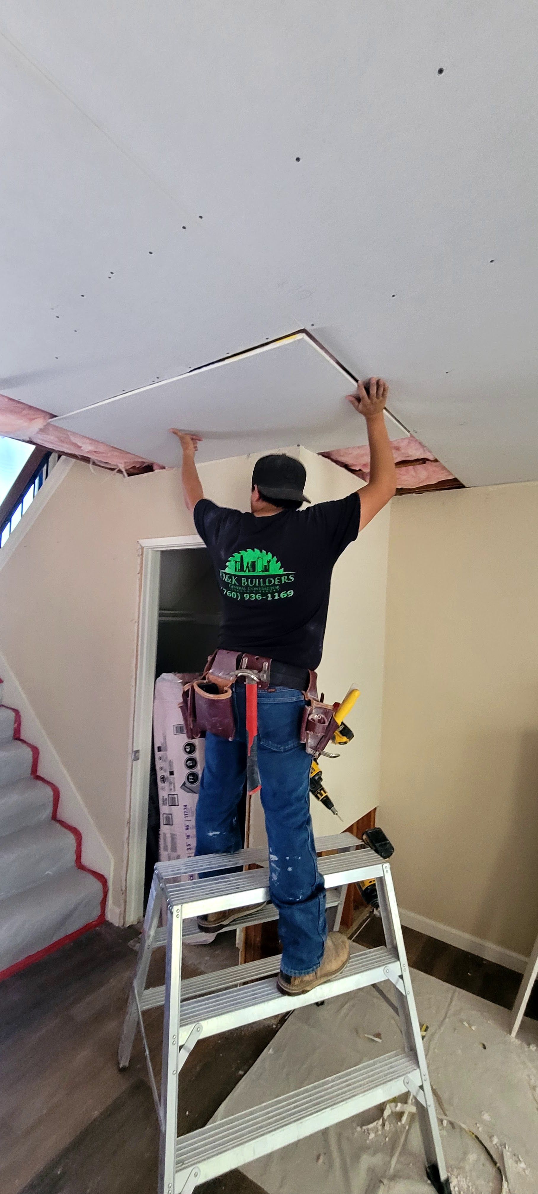 A person stands on a ladder, installing a rectangular ceiling panel into an opening in a residential interior.