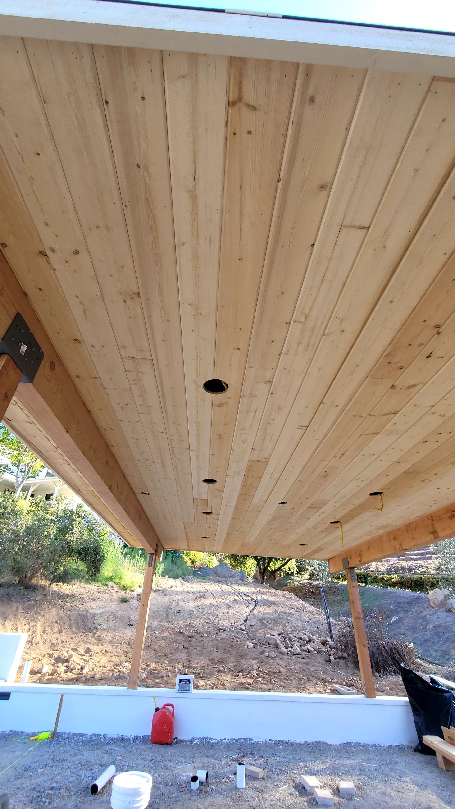 A low-angle view of a wooden ceiling structure under an unfinished outdoor canopy, with gravel ground visible below.