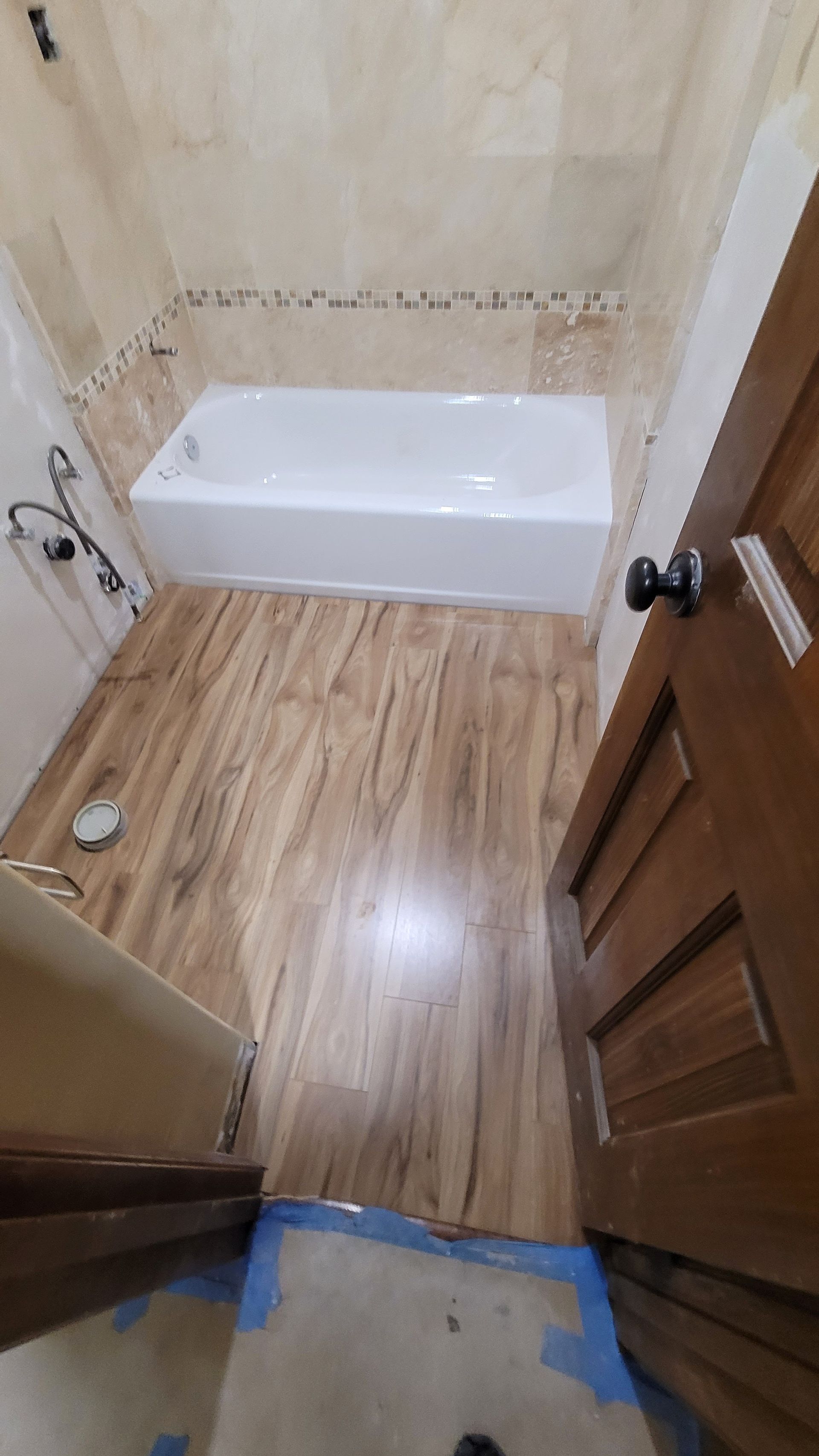 A bathroom with light wood-look flooring, a white bathtub, beige tiled walls, and a wooden door partially in view.