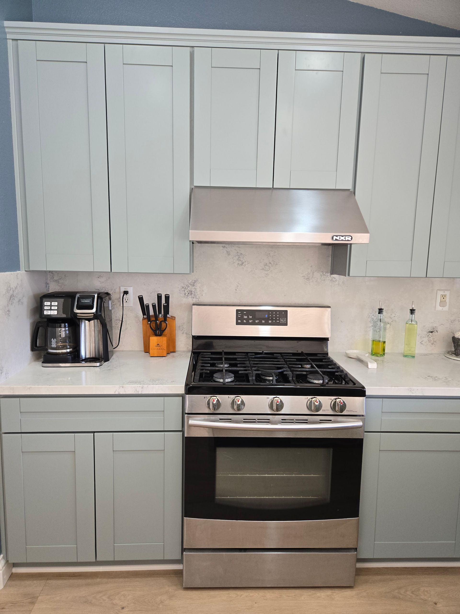 A kitchen scene featuring light blue cabinets, a stainless steel gas stove, a range hood, and a coffee maker on a counter.