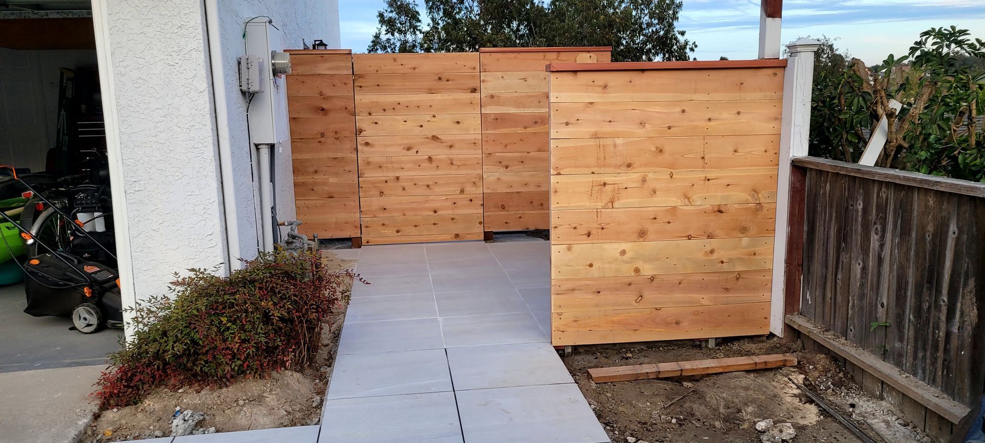 A paved walkway leads to a wooden privacy fence installed next to a white house exterior.