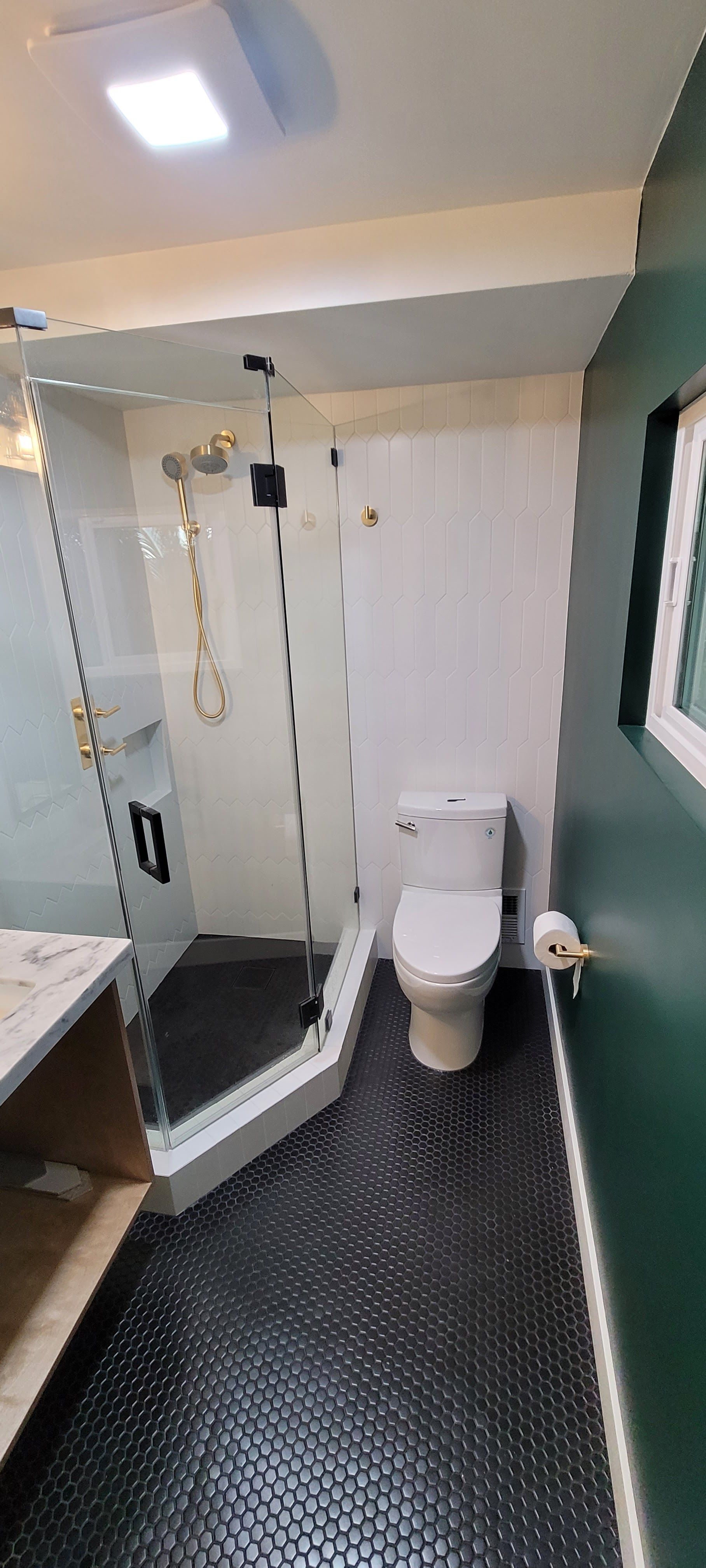 A bathroom with a corner glass shower, white toilet, and dark green walls featuring patterned black floor tiles.