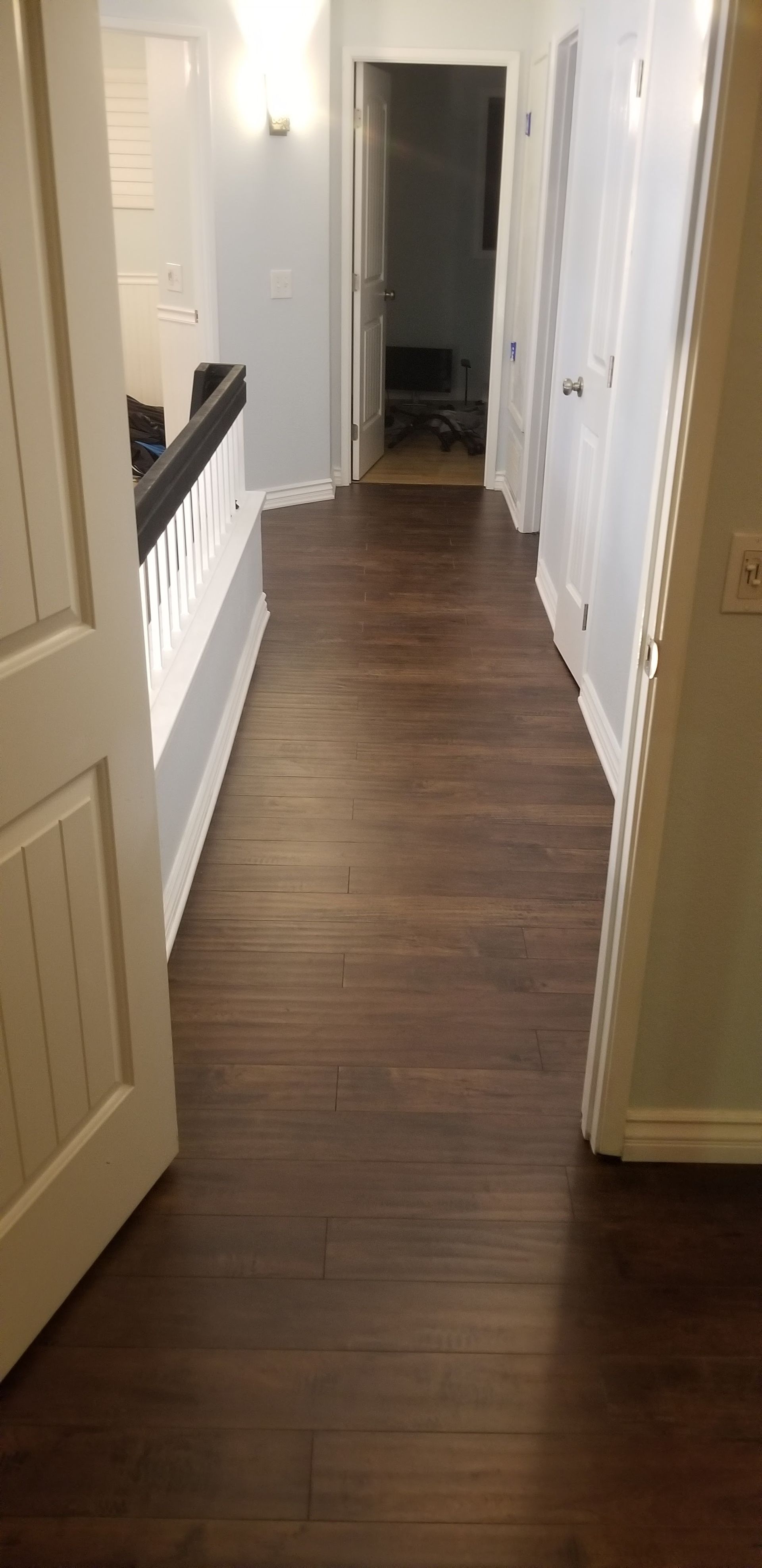 A view from a doorway looking down a hallway with dark wood-style flooring, white walls, and a staircase railing.