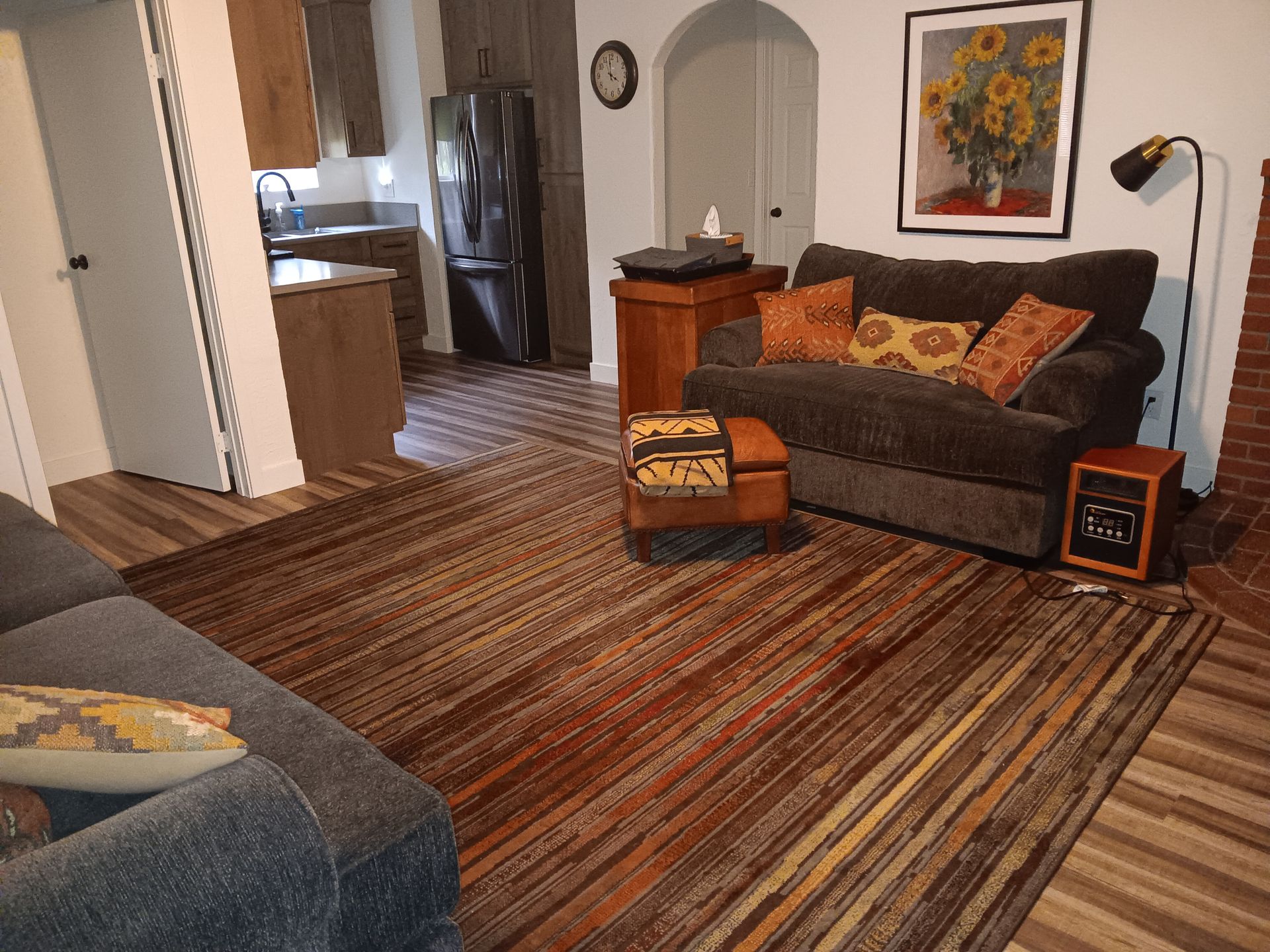 A living room featuring a striped rug, dark couch with patterned pillows, an ottoman, and a framed sunflower painting.
