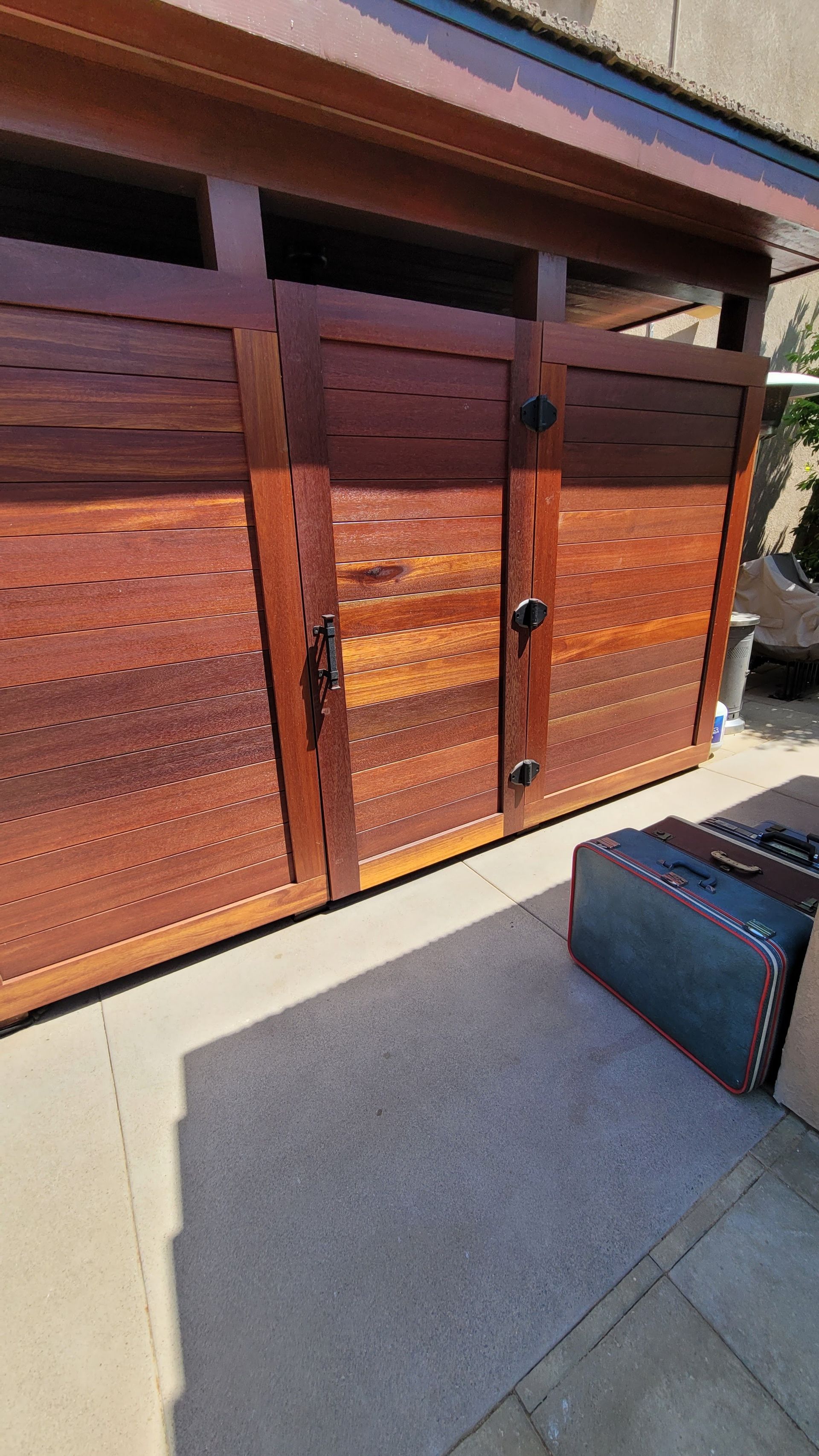 A wooden shed with horizontal slats and black hinges stands on a concrete patio next to a gray storage container.