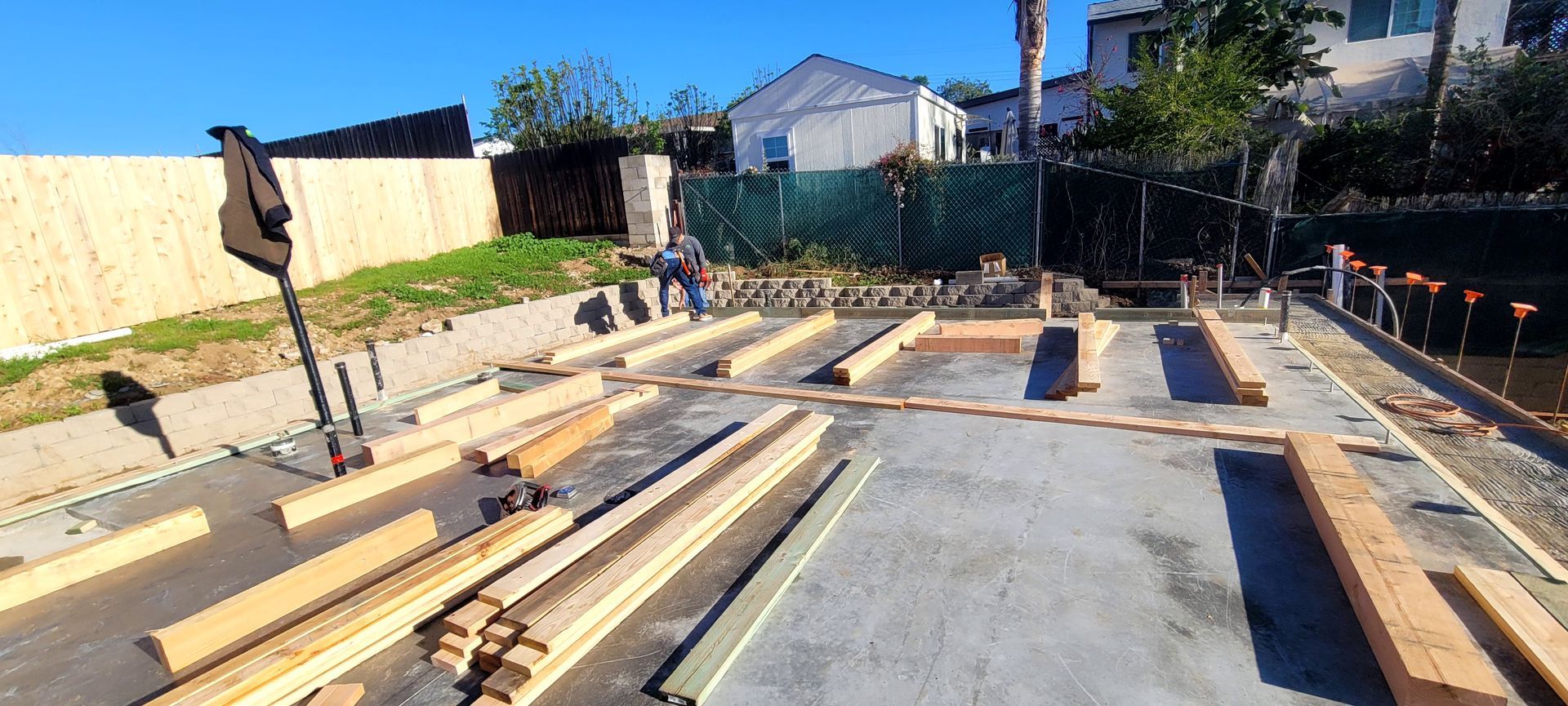 Wooden framing for a building foundation rests on a concrete slab in a construction site with a fence in the background.