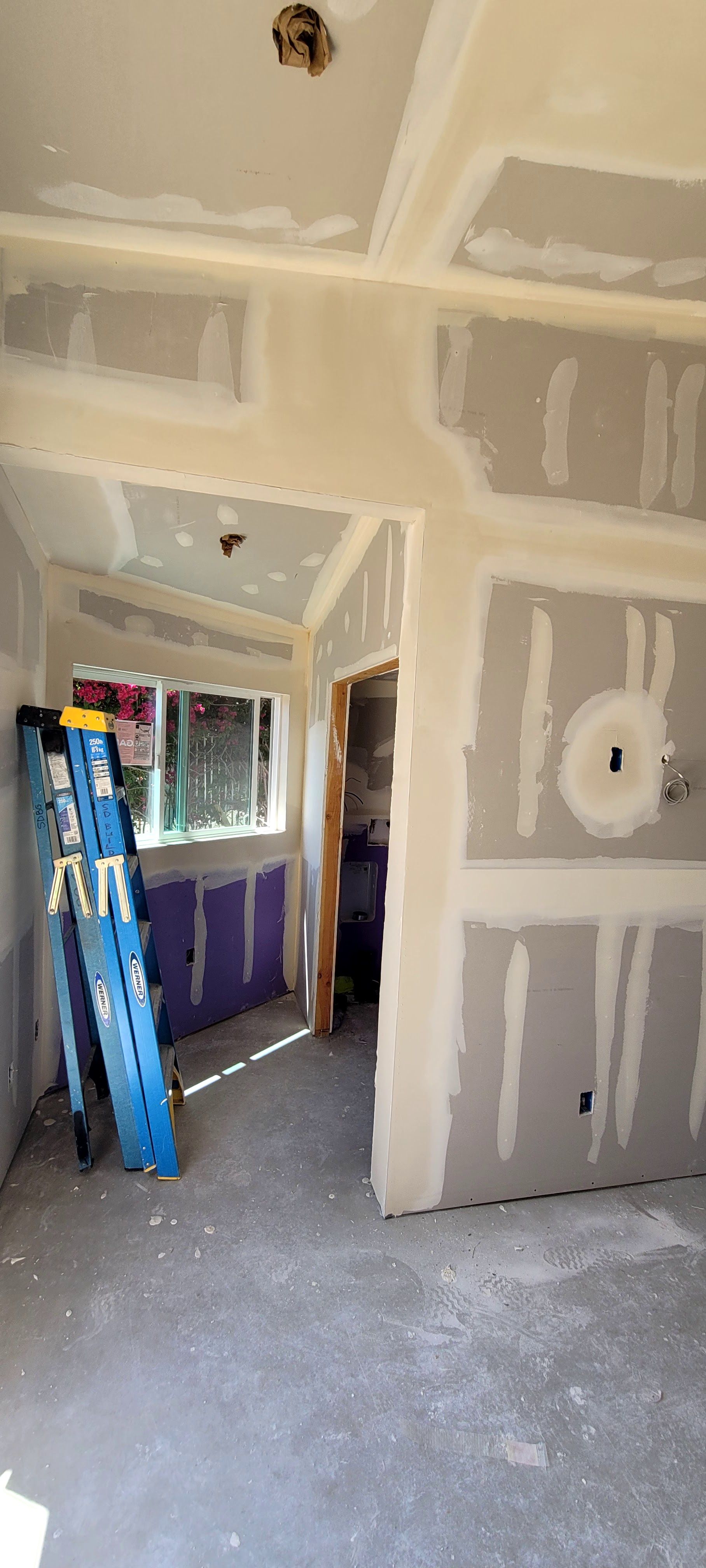 A room under renovation with taped drywall panels on walls and ceilings, a window with a blue ladder, and a door frame.