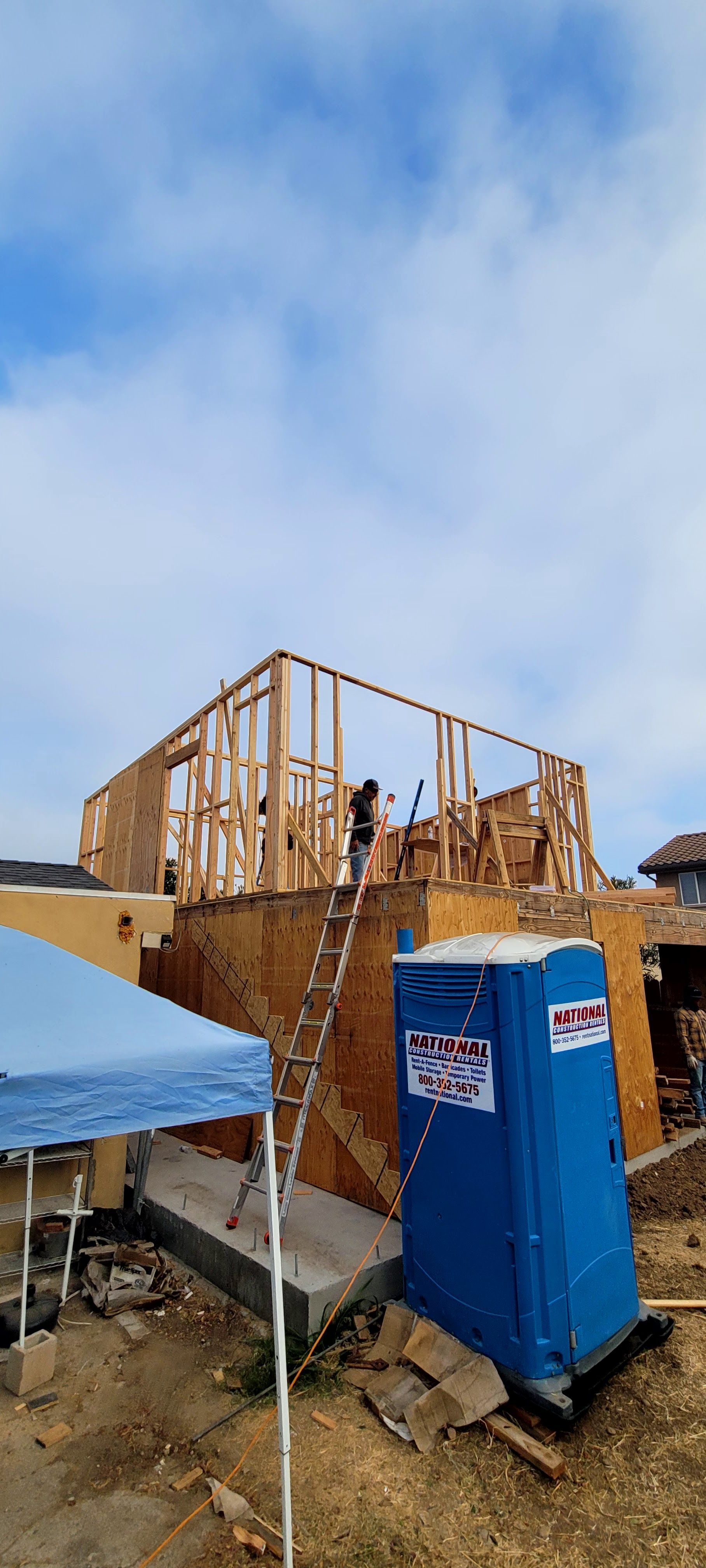 A person stands on a ladder next to the wooden wall frame of a house under construction with a blue portable toilet nearby.
