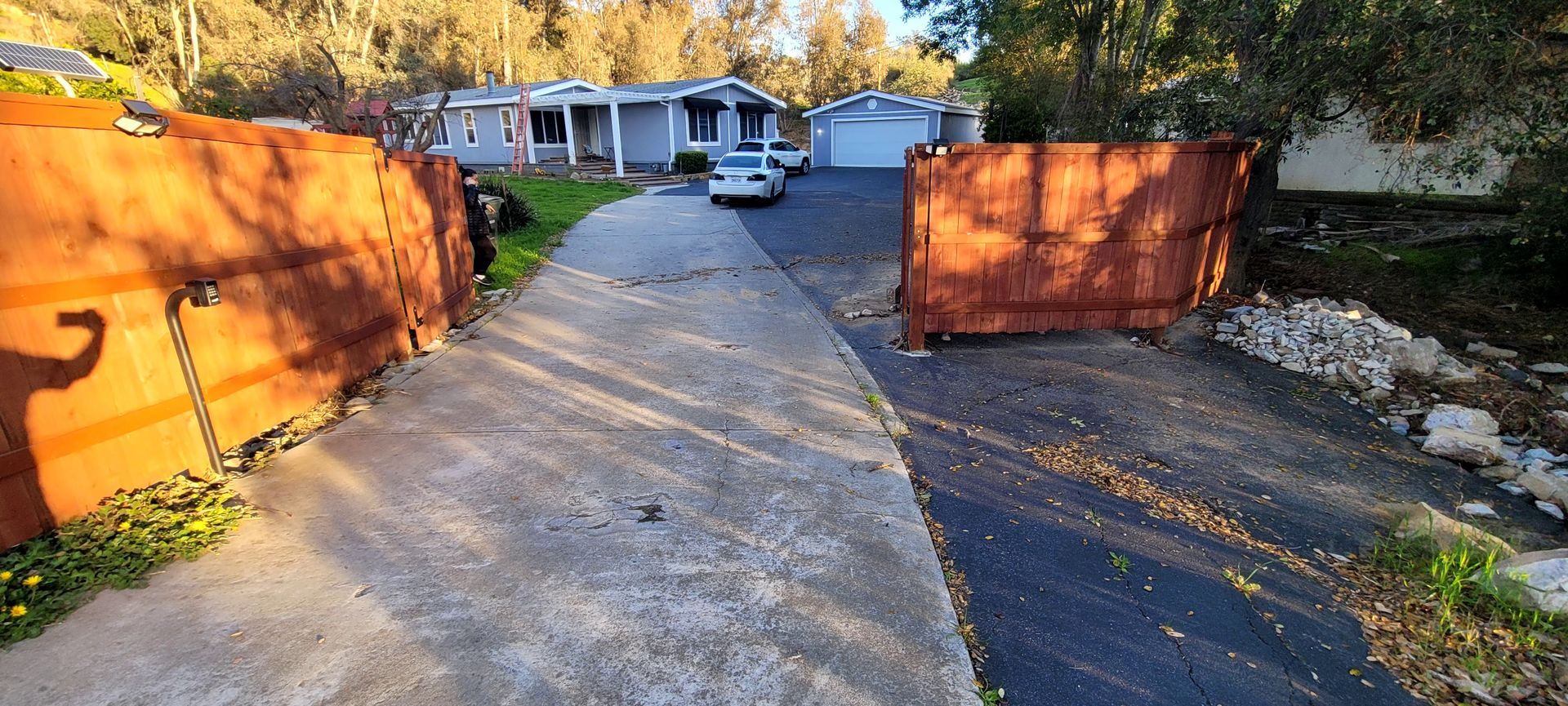 A driveway framed by two rusted metal walls leads to a house with a white garage door, with a car parked in the distance.