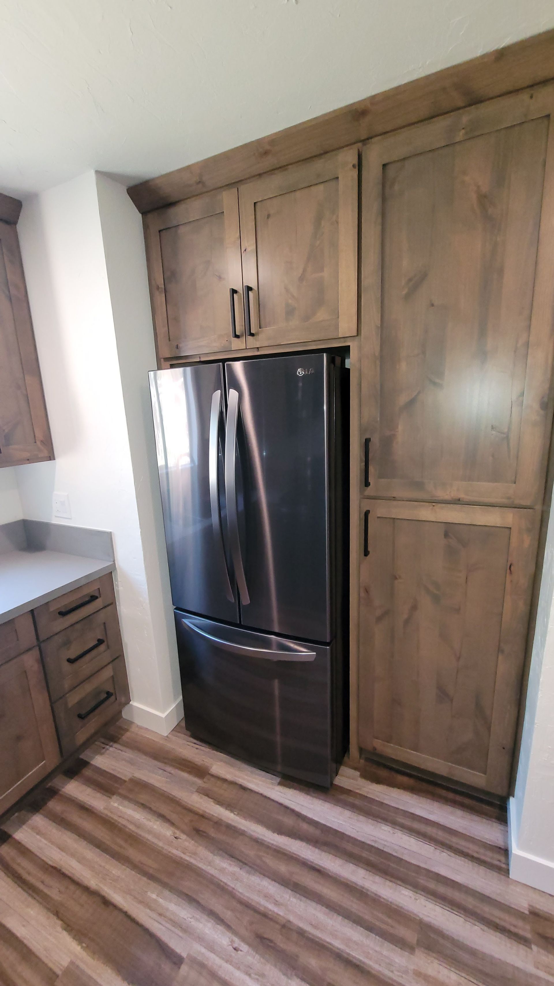 A stainless steel refrigerator fitted between wood-toned cabinets and a pantry in a kitchen with wood-look flooring.