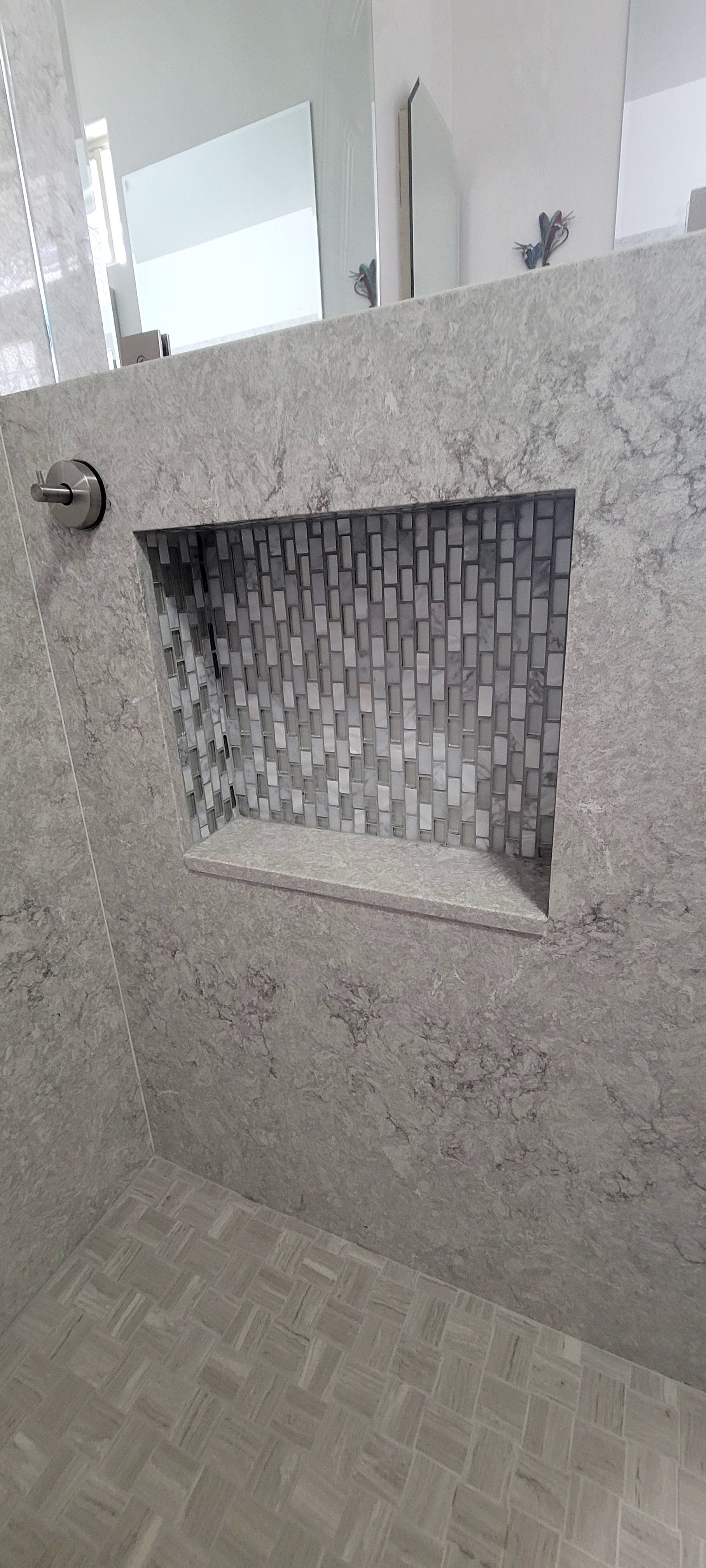 A shower wall with speckled grey stone tiling and a central rectangular recessed shelf lined with grey mosaic tiles.