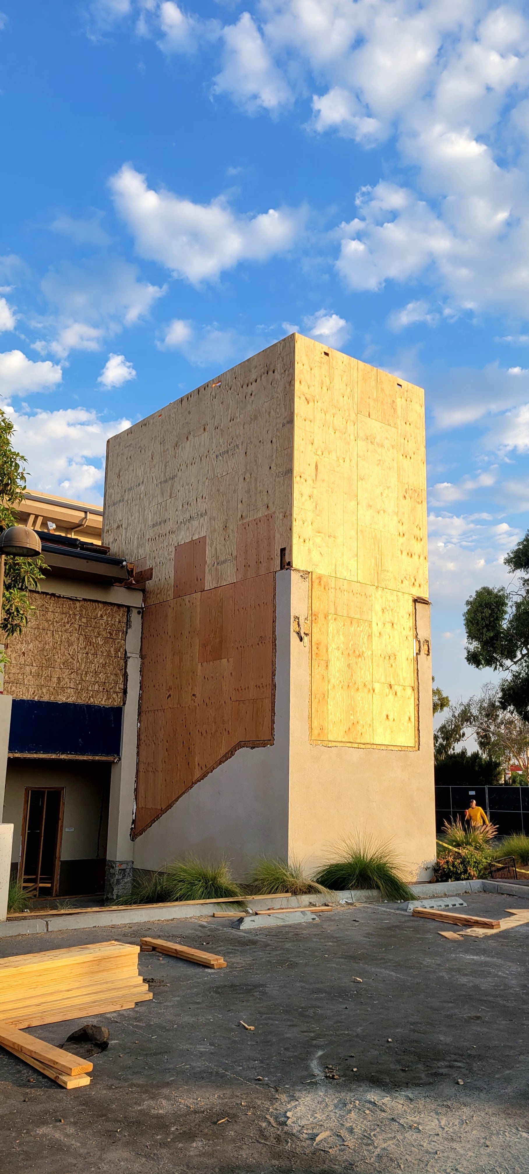 The exterior of a building undergoing construction, featuring exposed plywood walls and a tall, unfinished stucco tower.