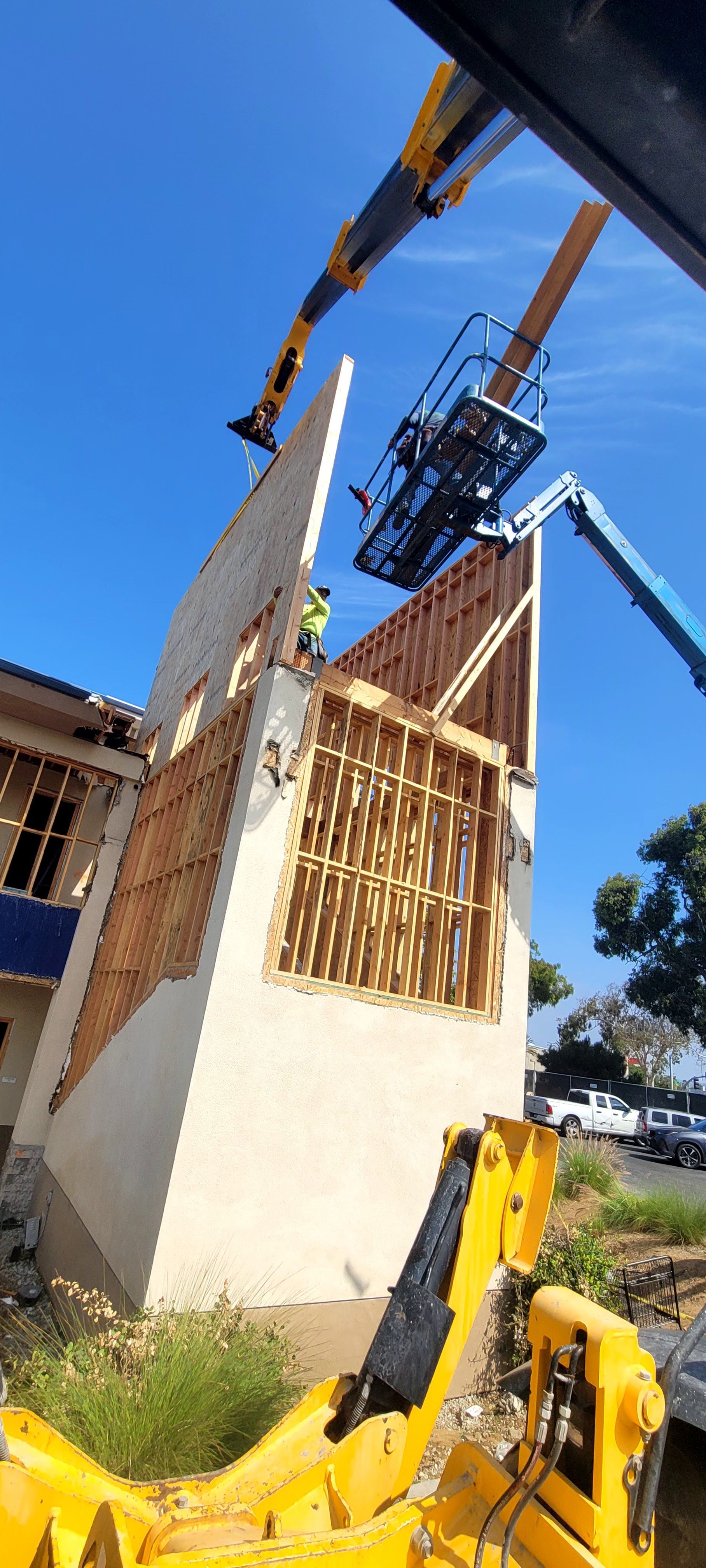 A construction worker in a high-reach lift works on the exposed wooden frame of a building exterior under a clear blue sky.