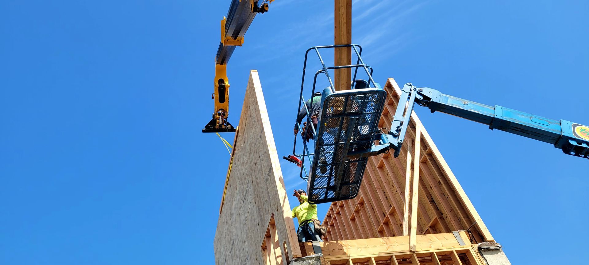 A construction worker in a high-visibility vest stands in a raised blue lift, installing a panel supported by a crane.