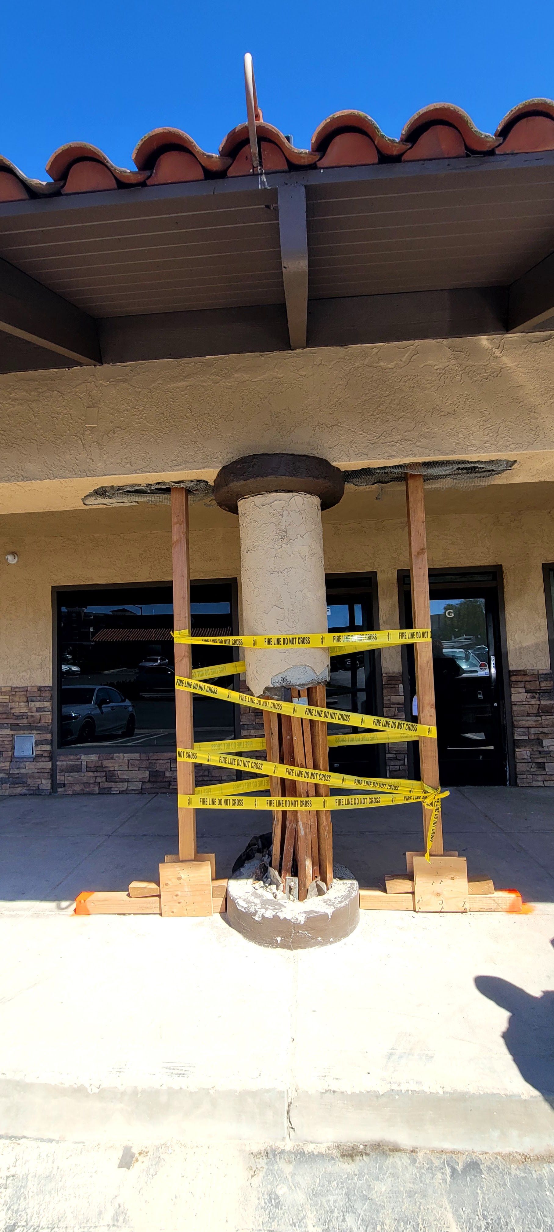 A damaged concrete support column, wrapped in caution tape and reinforced by temporary wooden shoring under a building roof.