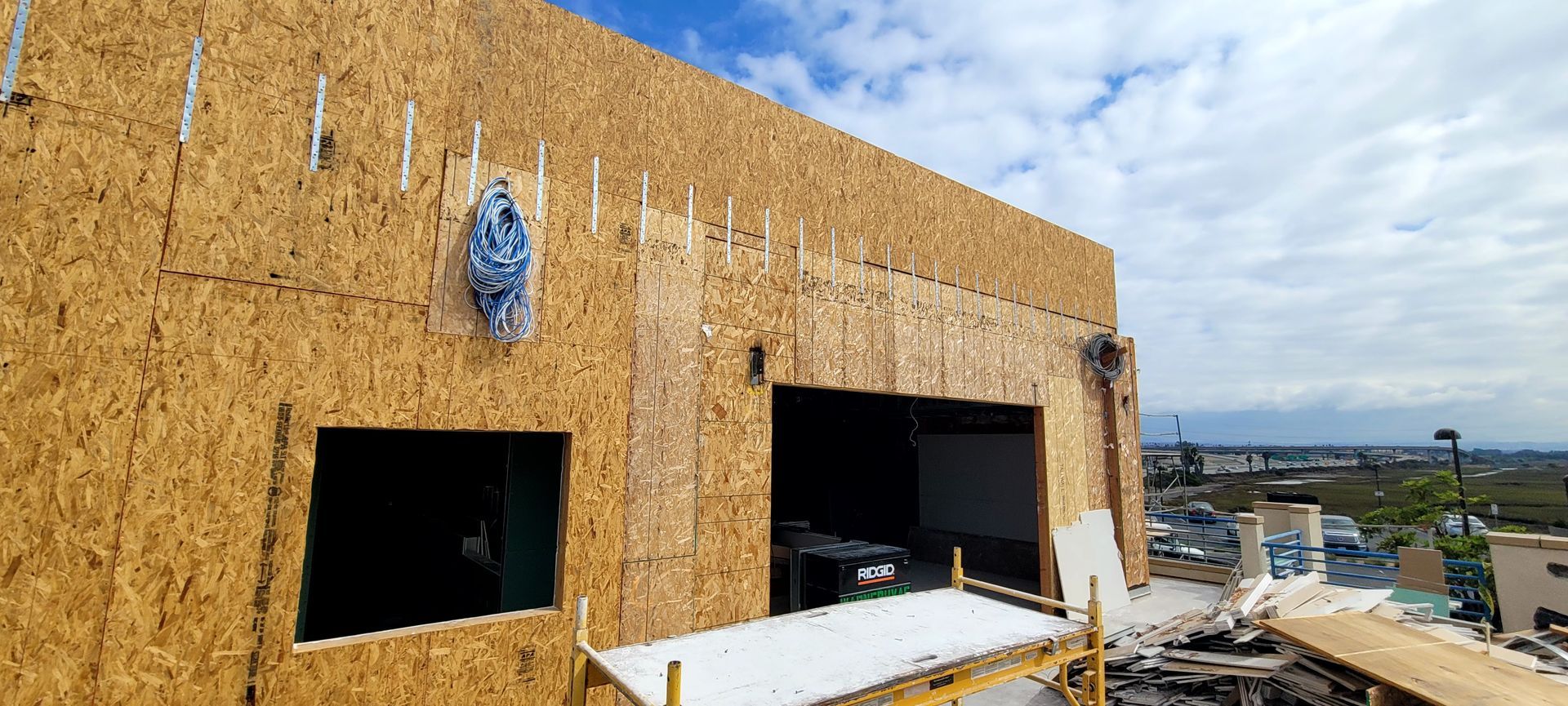 OSB-sheathed wall of a building under construction, featuring framed window and garage door openings against a blue sky.