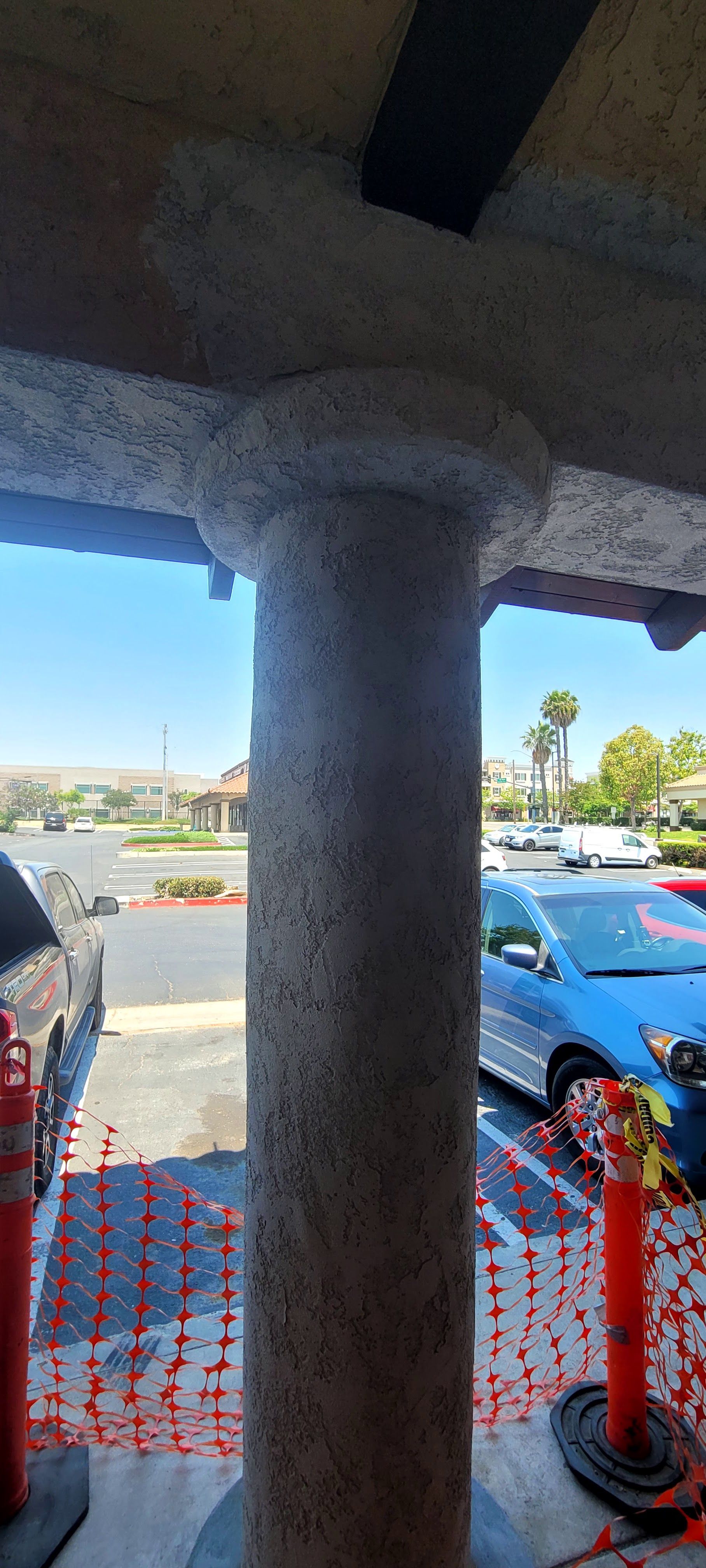 A textured, light-colored concrete pillar stands in front of a parking lot with several cars and safety barriers.