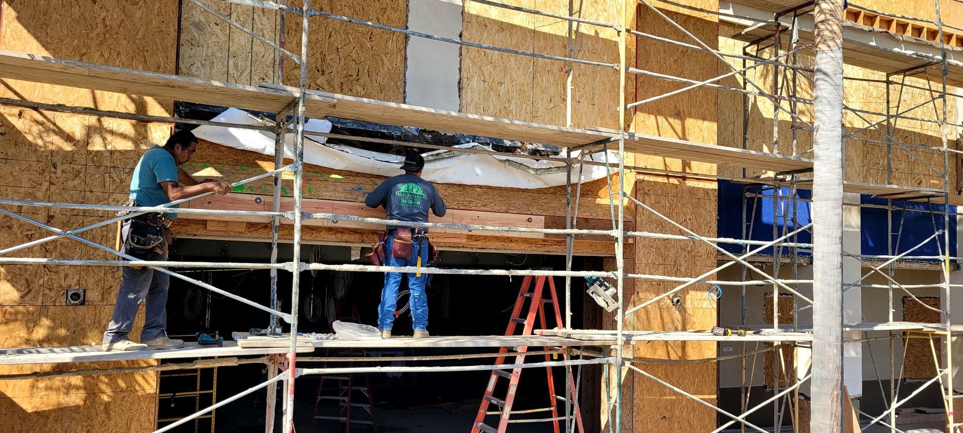 Two construction workers on scaffolding install a header beam over a large open doorway in a framed building exterior.