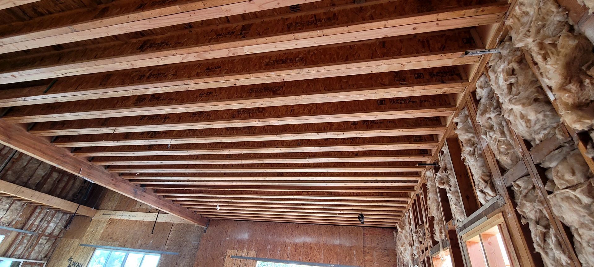 Unfinished ceiling with exposed wooden floor joists and a wall partially insulated with fiberglass batts.